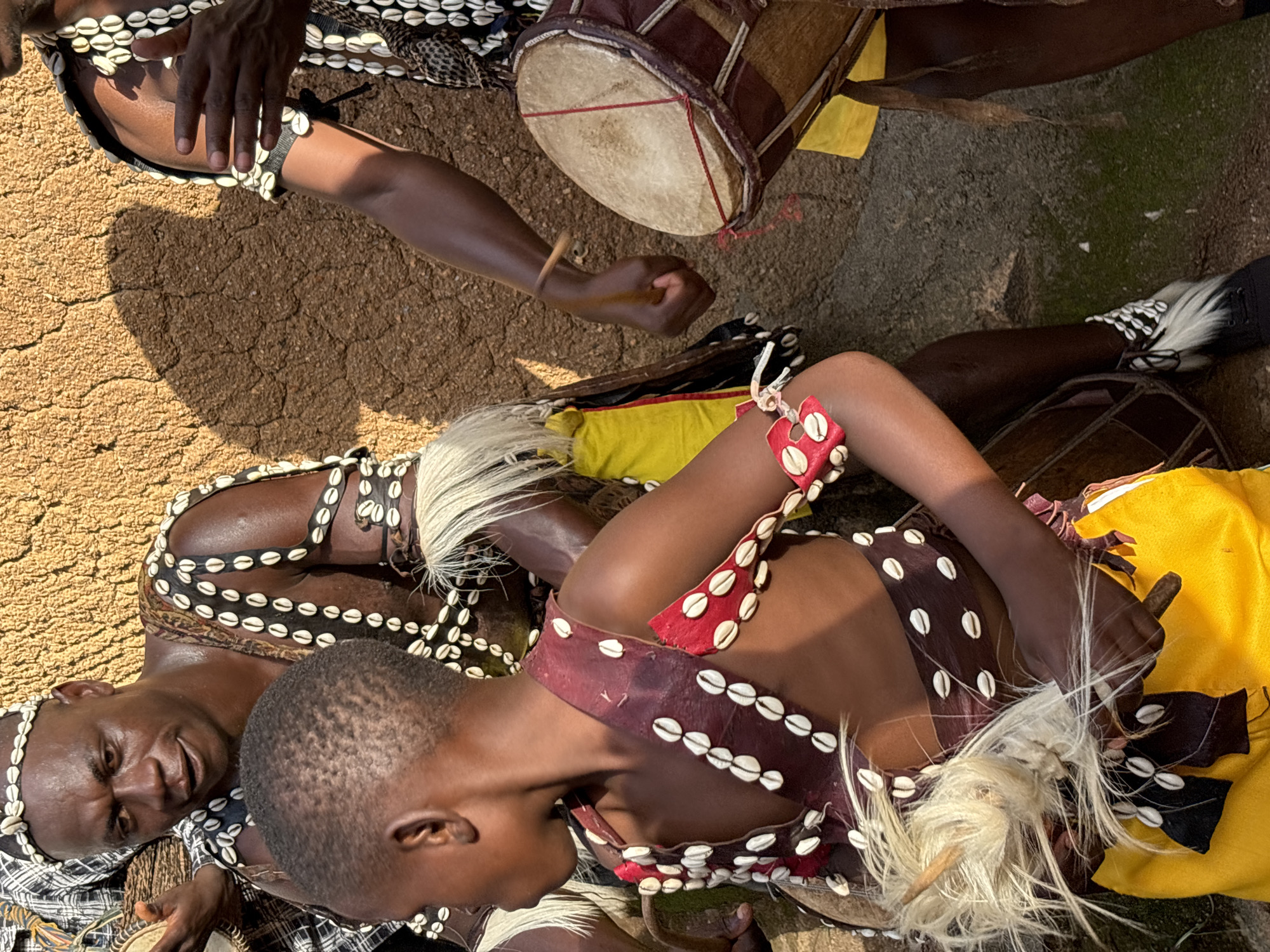 Two people are engaged in a traditional performance, likely a dance or ritual, in Jos, Nigeria. They are adorned in elaborate costumes featuring cowrie shells, animal hide, and white feathery adornments. One individual, an adult male with a warm smile, wears a headdress and necklace made of cowrie shells, along with a patterned fabric around his head and shoulders. He is playing a small drum.
The second individual, a younger person, is lying down, also adorned in a similar fashion with cowrie shells and feathery elements. They wear a red and brown vest decorated with cowrie shells, and what appears to be a yellow garment underneath. Their head is resting on their arm, and their body is positioned in a relaxed, almost contemplative pose, contrasting with the active stance of the drummer.
In the background, another drum, larger and made of leather, rests on a textured, earthy surface, possibly a dirt or stone ground. The lighting suggests it is daytime, with warm sunlight casting shadows. The overall scene evokes a sense of cultural celebration and tradition, with the performers showcasing their heritage through music and adornment. No visible text is present in the image.