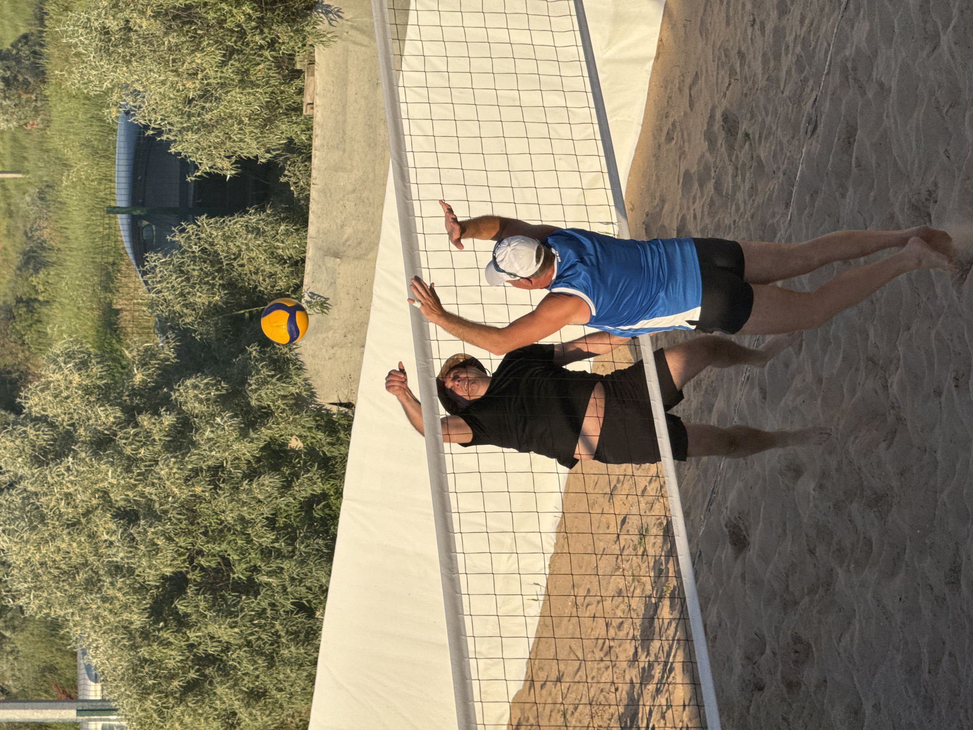 Two men are playing beach volleyball on a sandy court. The man on the left, wearing a black t-shirt and black shorts, is in mid-air, reaching up to hit the ball. He has a straw hat on his head. The man on the right, wearing a blue tank top with white accents and black shorts, is also reaching up, seemingly to block the hit. He has a white cap on his head. A volleyball, blue and yellow, is in the air between them, above the net. The net is strung across the court. In the background, there is a white structure, a line of green bushes, and beyond that, a fence and a dark car. The scene appears to be outdoors, possibly in the city of Shchelkino, Ukraine, given the reference provided. The ground is covered in sand, and shadows are cast by the players and the net. The lighting suggests it is late afternoon or early evening.