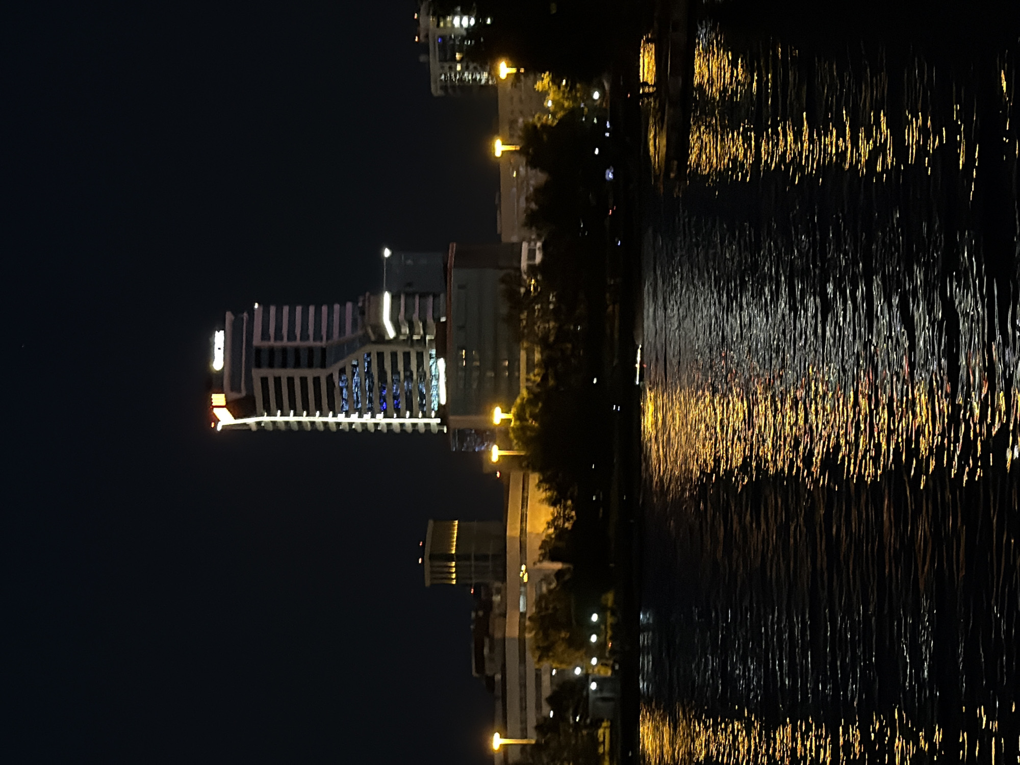This is a nighttime photograph of the Yekaterinburg City Pond, showcasing a cityscape illuminated by artificial lights. A prominent skyscraper with a distinctive illuminated top and a logo that appears to read "BISHOP" stands tall in the background, dominating the skyline. To its left, other buildings with illuminated windows and structures are visible. The foreground is occupied by the dark water of the pond, reflecting the bright yellow and orange lights of the city, creating shimmering patterns on the surface. Trees and streetlights line the banks of the pond, adding to the ambient illumination. The overall scene depicts a vibrant urban environment at night, with the water serving as a reflective canvas for the city's glow.