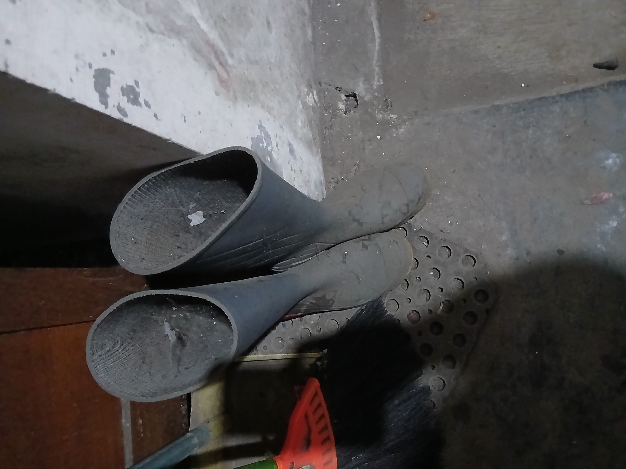 In a dimly lit, possibly indoor storage area, likely in Tenggarang, Indonesia, two tall, dark rubber boots stand upright on a patterned mat. To the left, the bristles and orange handle of a broom are visible, suggesting a cleaning or storage space. The boots appear dusty, indicating they have been used and placed there for a period. The surrounding environment is utilitarian, with rough concrete walls and what appears to be wooden paneling or furniture in the background. The overall impression is one of a functional, perhaps neglected, area.