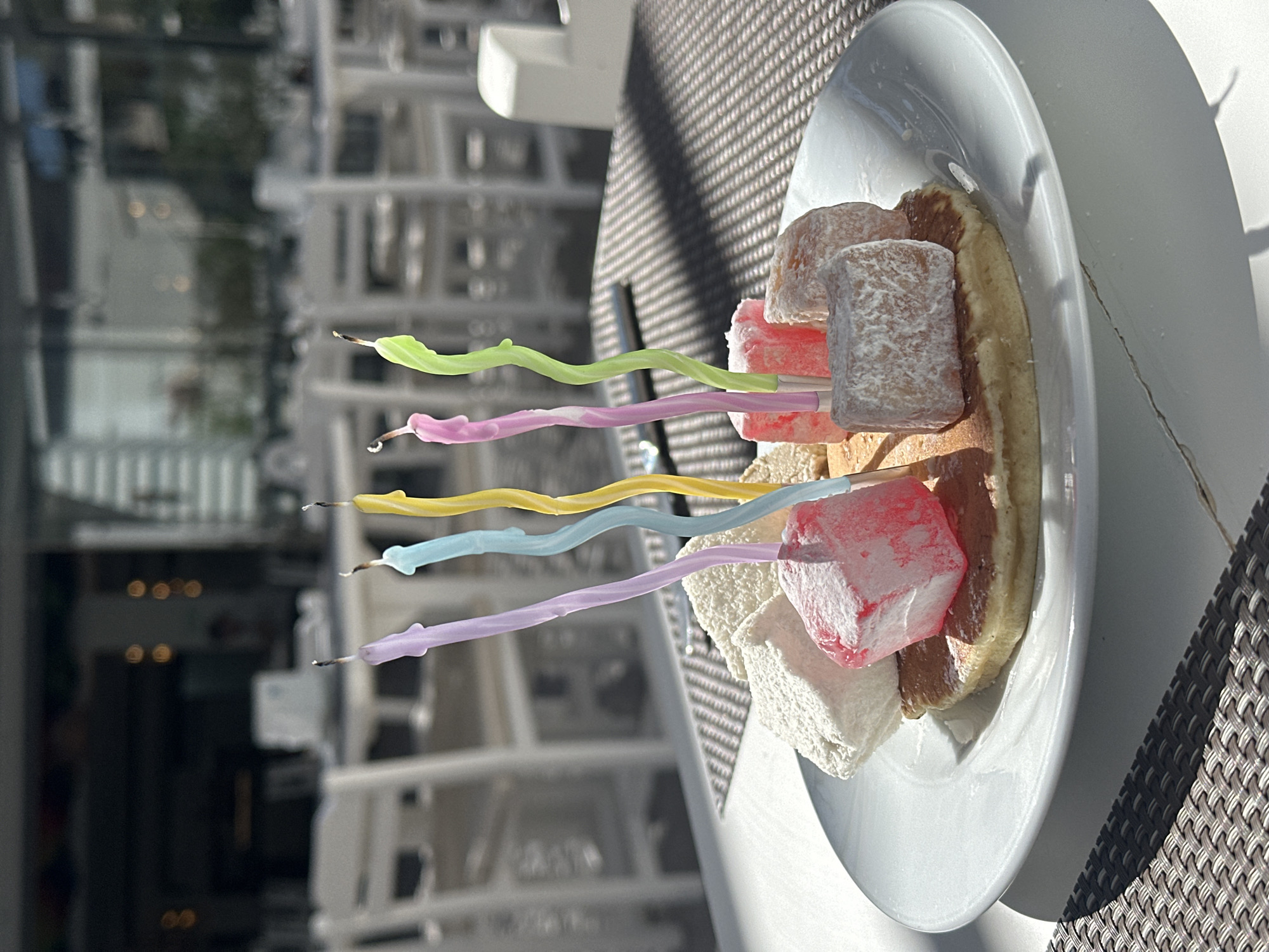 The image shows a white plate with food, likely Turkish delight, and five unlit birthday candles, set on a table with a woven surface. The background features an out-of-focus outdoor setting with architectural structures, suggesting an urban environment. Based on the context clues and the presence of Turkish delight, the location is possibly in Fethiye, Turkey. There are no visible people or explicit interactions. The objects present are the plate, the Turkish delight, the birthday candles, and the table. The environment is sunny and outdoors, with some blurred architecture in the backdrop.