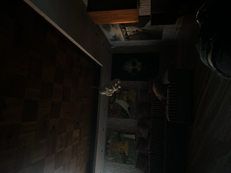 A room features a dark, wooden-paneled wall. Several framed images hang on an adjacent wall, including one with a prominent white hand with a red ring and another with a face. A white sculpture is mounted on the wall. A striped sofa sits alongside the art. Lighting in the room is dim.