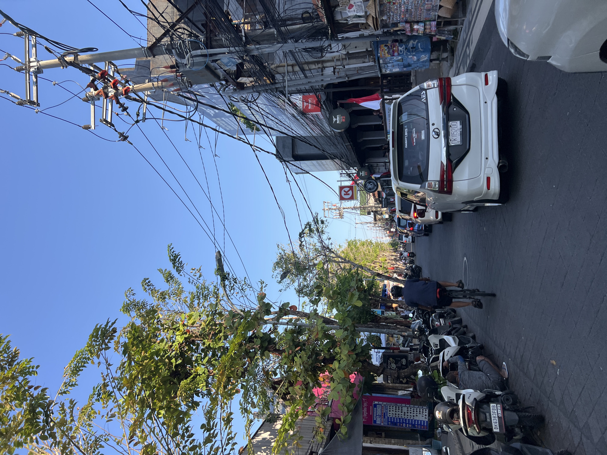 The image depicts a street scene in Kuta, Indonesia. The street is paved and lined with buildings and trees. Several motorcycles are parked on the side of the road. A person is riding a bicycle in the street, and a white van is driving in the same direction. There are many power lines overhead, creating a dense network. The buildings appear to be commercial establishments, with signs and storefronts visible. The sky is clear and blue. The overall impression is of a busy urban environment with a lot of activity.