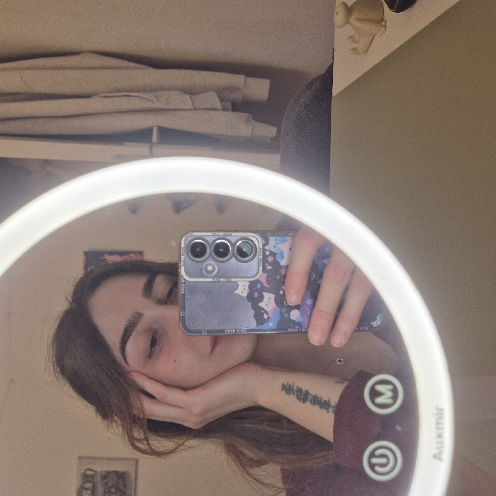 An individual, appearing to be female, with long brown hair, is depicted taking a selfie reflection in a circular, illuminated Auxmir brand makeup mirror within an indoor setting in Strasbourg, France. The person is wearing a dark reddish-maroon long-sleeved top, and a black text tattoo is visible on their left inner forearm. Their left hand rests against their cheek, while their right hand holds a smartphone with a distinctive case featuring cartoon cats and stars. The smartphone's multi-lens camera array is clearly visible. Reflected in the background are various room elements: a framed image on a light-colored wall to the left, and clothing (likely a light-colored hooded garment) hanging on a wall to the right. A light green-beige surface, presumably a desk or vanity, is visible in the lower right, holding a small light-colored figurine and another dark object. Two illuminated touch controls, power and mode, are visible on the bottom rim of the mirror.