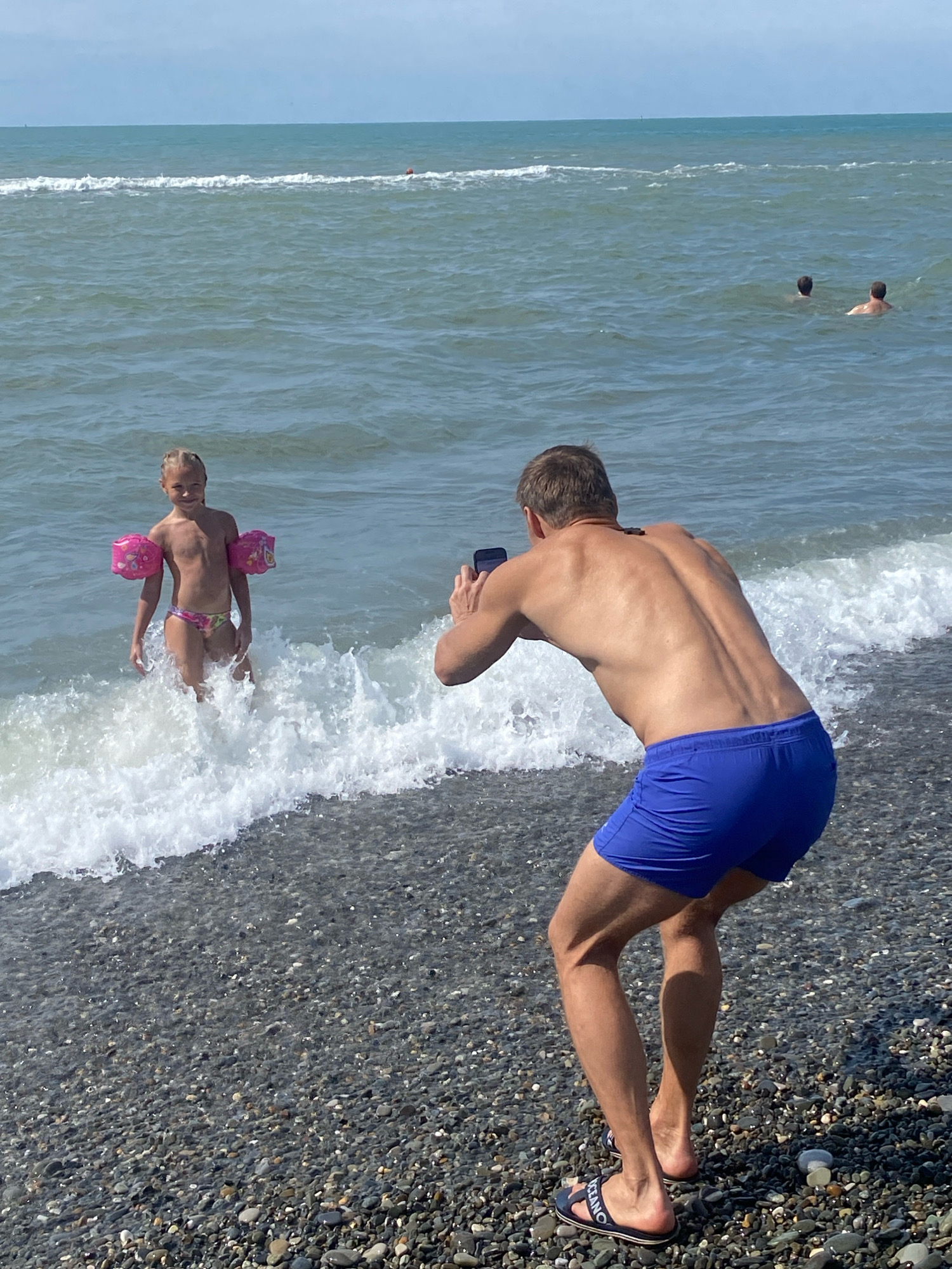 The media file depicts a lively beach scene in Sochi, Russia, under a bright, sunny sky. The water is a deep blue with white-capped waves crashing onto a pebble-strewn shore.
The primary subjects are a young girl and a man. The girl, standing in the shallows with waves swirling around her, wears a colorful bikini and pink arm floaties, suggesting she is enjoying a day of swimming or playing in the sea. She has blonde hair styled in braids and is looking towards the viewer with a slight smile.
Behind her, a man is captured in the act of taking a photo with his phone. He is wearing blue swim trunks and is bent slightly forward, focused on framing his shot. His muscular back is visible, and he is wearing dark sandals. In the distance, two other men can be seen swimming in the sea.
The scene evokes a sense of a pleasant summer day at the beach, filled with recreational activity. The overall mood is relaxed and joyful, typical of a vacation or a day out with family. The time of day appears to be midday, given the strong sunlight casting shadows. The location is clearly identifiable as Sochi, Russia, indicated by the provided context. There is no visible text in the media file itself.