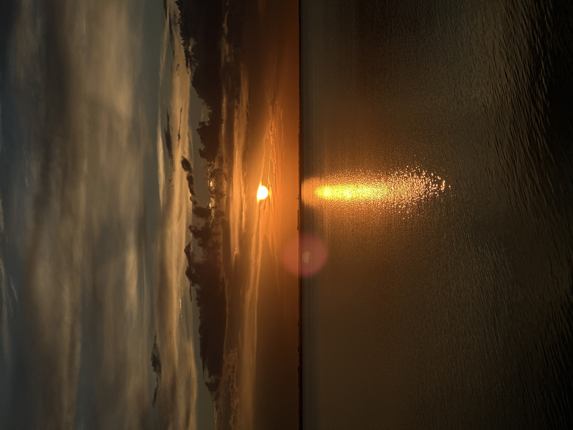 The image depicts a sunset over a body of water, likely a lake or sea, with a distant shoreline visible. The sky is filled with dramatic clouds, ranging in color from dark gray and brown to vibrant orange and yellow near the horizon where the sun is setting. The sun itself is a bright, golden orb, partially obscured by clouds, casting a shimmering reflection on the water's surface. The water is dark and calm, with small ripples. On the distant shoreline, there appear to be some industrial structures or buildings silhouetted against the bright sky. The overall mood of the image is serene and majestic, characteristic of a sunset. The provided crops highlight the textured clouds and the reflective qualities of the water. No people or specific landmarks that would definitively identify the location as Kokshetau, Kazakhstan, are clearly visible.