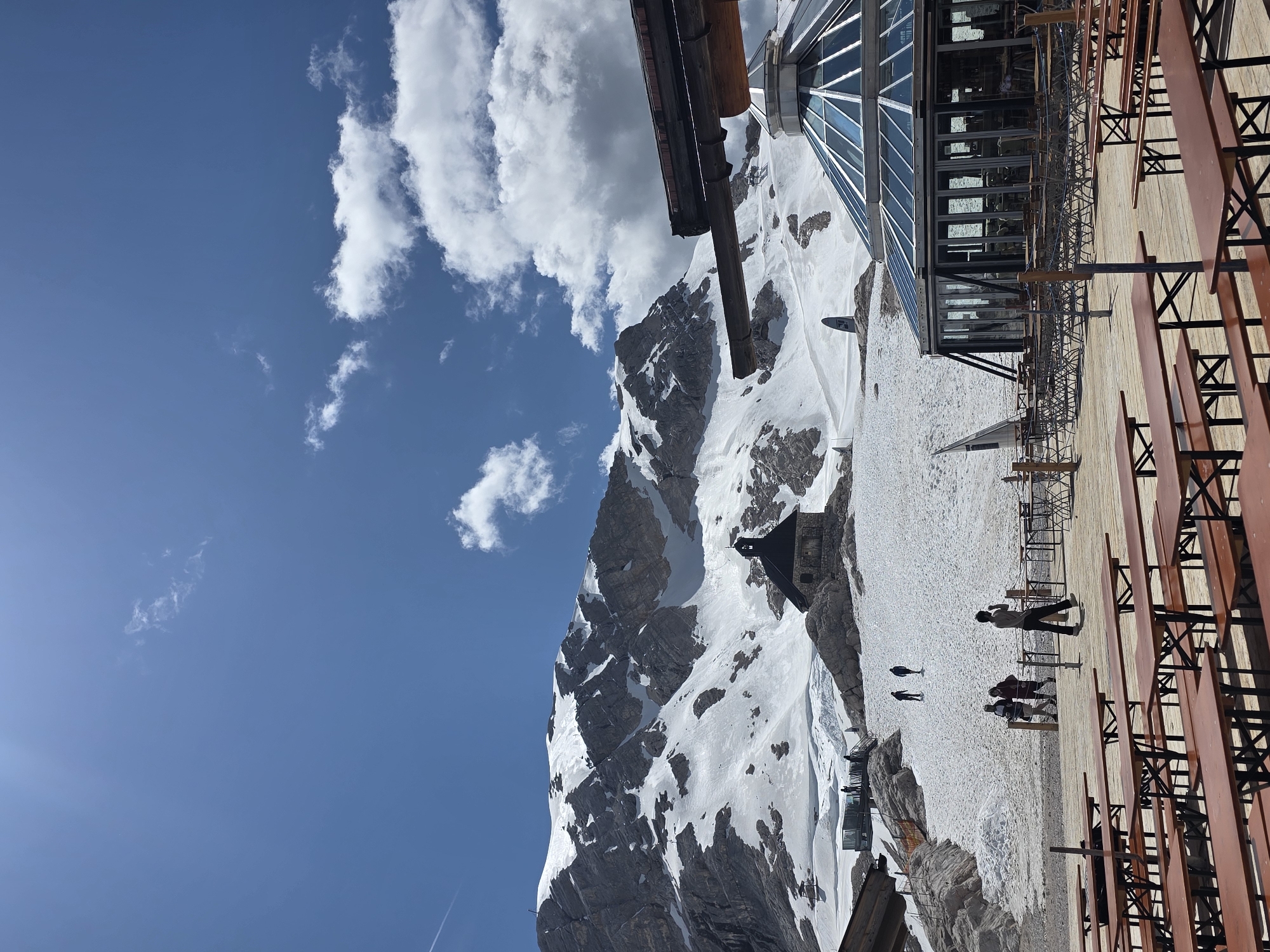 A bright, sunlit, wide-angle photograph captures a mountain landscape under a blue sky with scattered white clouds. In the foreground, a wooden deck is set with numerous long, narrow picnic tables and benches. Beyond the deck, a circular, glass-enclosed building with a domed roof is visible, with outdoor seating arranged in front of it. Further into the scene, a small stone building with a dark roof and a bell tower sits on a snow-covered slope. A large, snow-capped mountain range dominates the background. Several people are scattered across the snowy area, some walking and others appearing to stand. A thin white contrail is visible in the upper left corner of the sky. The scene suggests a tourist destination in a mountainous region, likely at a high elevation given the snow.