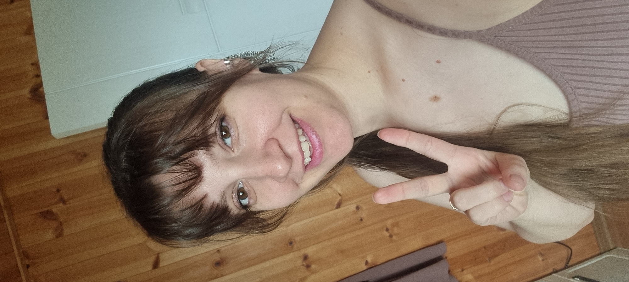 A woman is shown in a selfie, smiling and holding up a peace sign. She has brown hair with bangs and is wearing a light purple top. She has a ring on her finger, and an earring in her left ear. The background includes a light-colored wall and a wooden floor. There is no evidence to suggest the location is Dunnae-myeon, South Korea.
