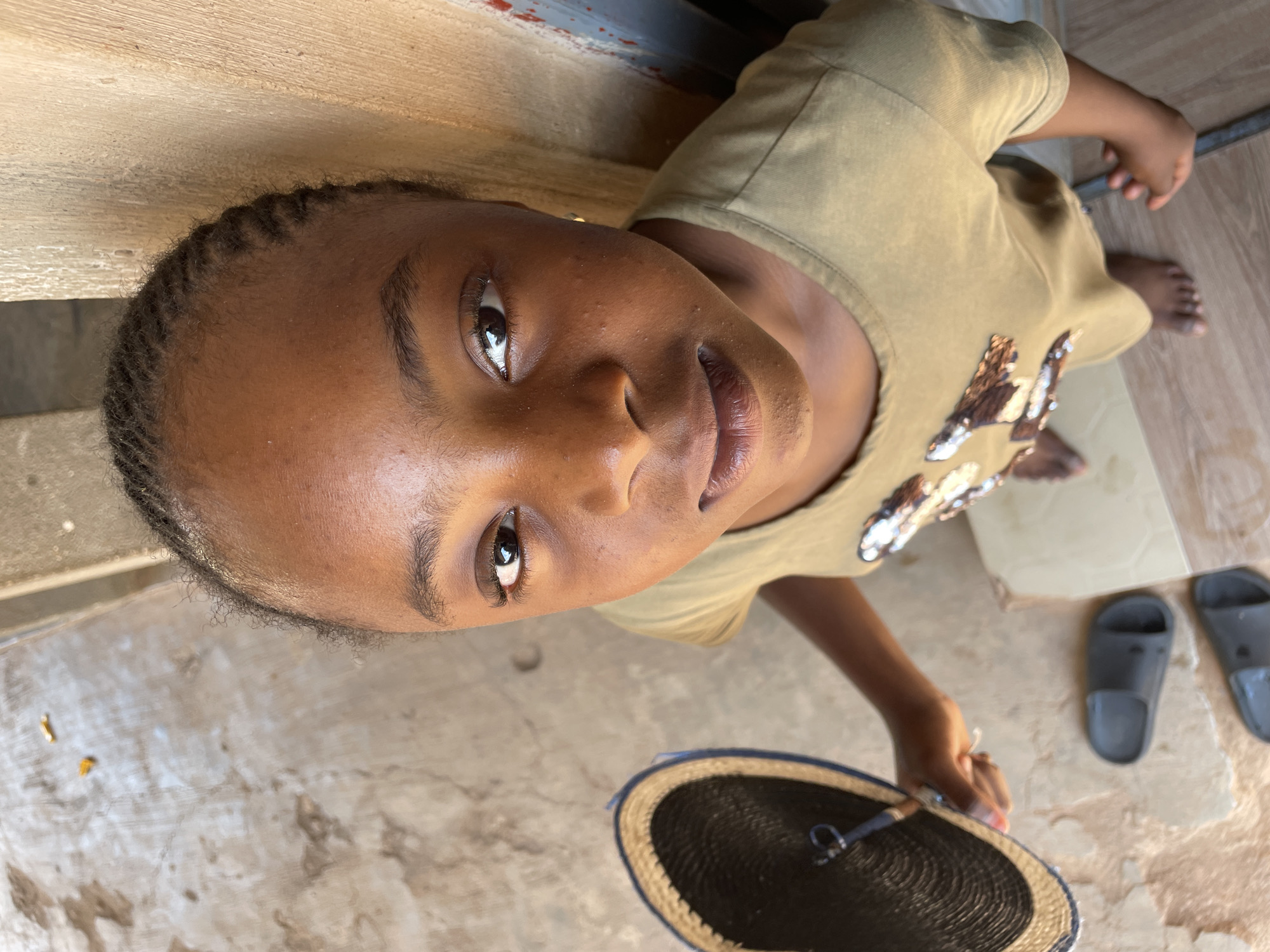 A young girl with dark skin and braided hair looks up at the camera, holding a round woven object with a dark center and light-colored rim. She wears a tan shirt with sequin-like decorations. The background appears to be an outdoor area, possibly a patio or courtyard. The ground is textured with a worn, concrete-like surface. A pair of gray rubber sandals are visible in the background. The location is Jos, Nigeria.