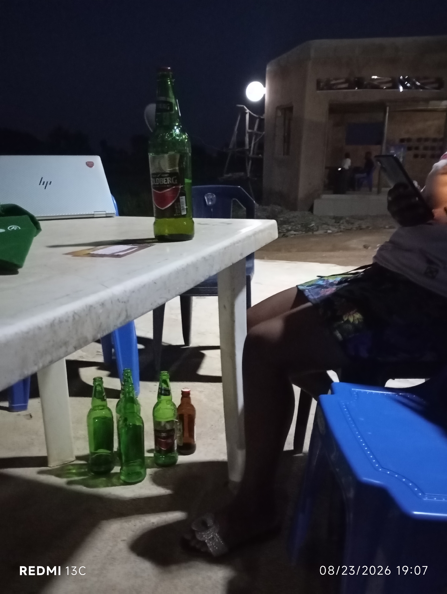 In Shendam, Nigeria, at night, a group of people appear to be socializing around a plastic table. A laptop, possibly a HP, is open on the table. Several bottles of beer, one of which is labeled "GOLDBERG", are also on the table and on the ground, suggesting a casual gathering. A person, whose legs and torso are visible, is seated in a blue chair and holding a mobile phone. In the background, a building with an open structure and a light fixture suggests a commercial or residential area. The overall scene conveys a sense of relaxed evening activity.