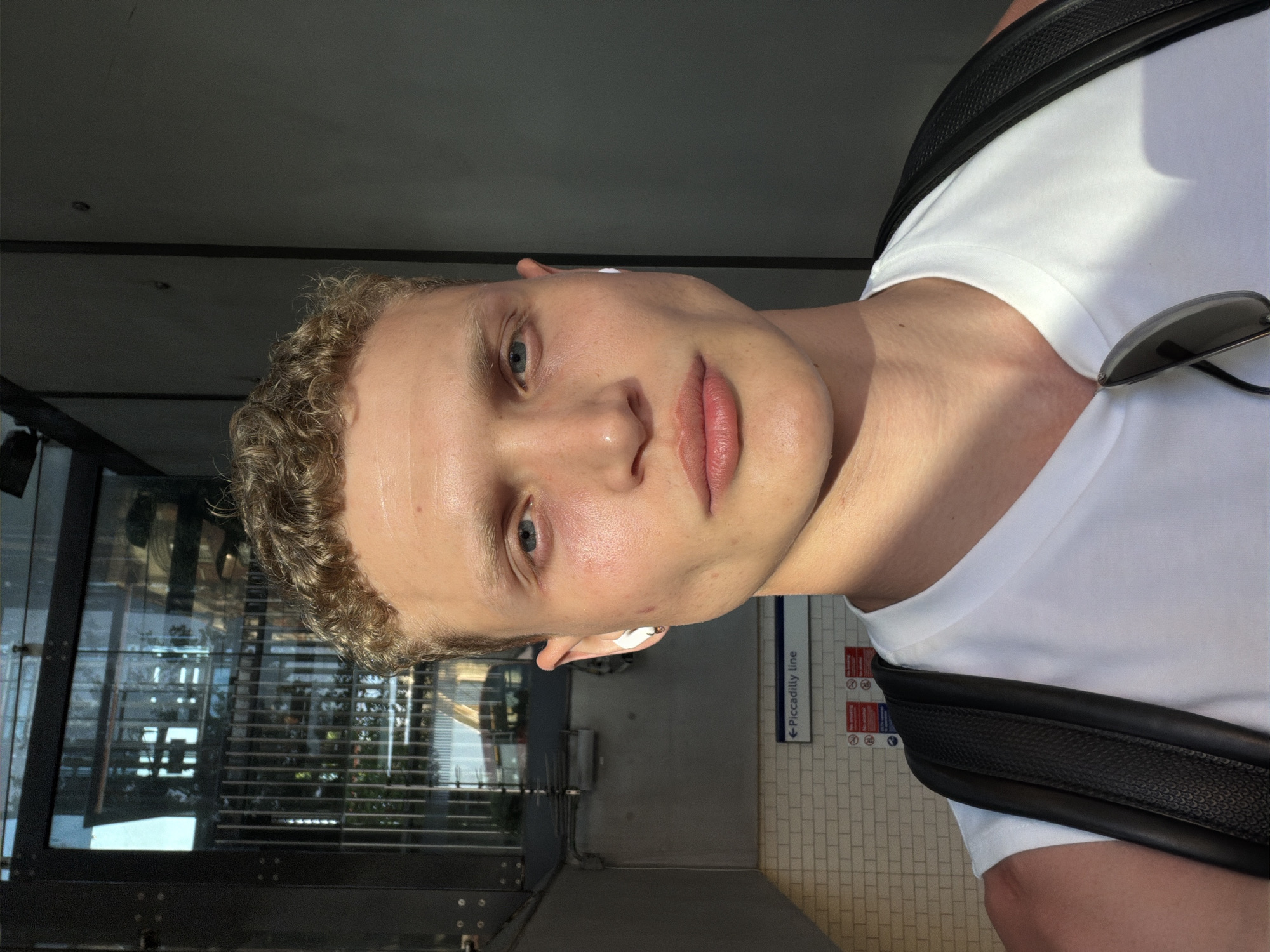 A Caucasian man is positioned in a well-lit underground station, which can be determined from the "Piccadilly Line" sign, indicating a location in London, United Kingdom. He has fair skin, curly blond hair, and blue eyes, and is wearing white wireless earbuds. He is wearing a white T-shirt and a black backpack. A pair of sunglasses is hooked onto his shirt. The background shows a station platform with tiling and glass partitions.