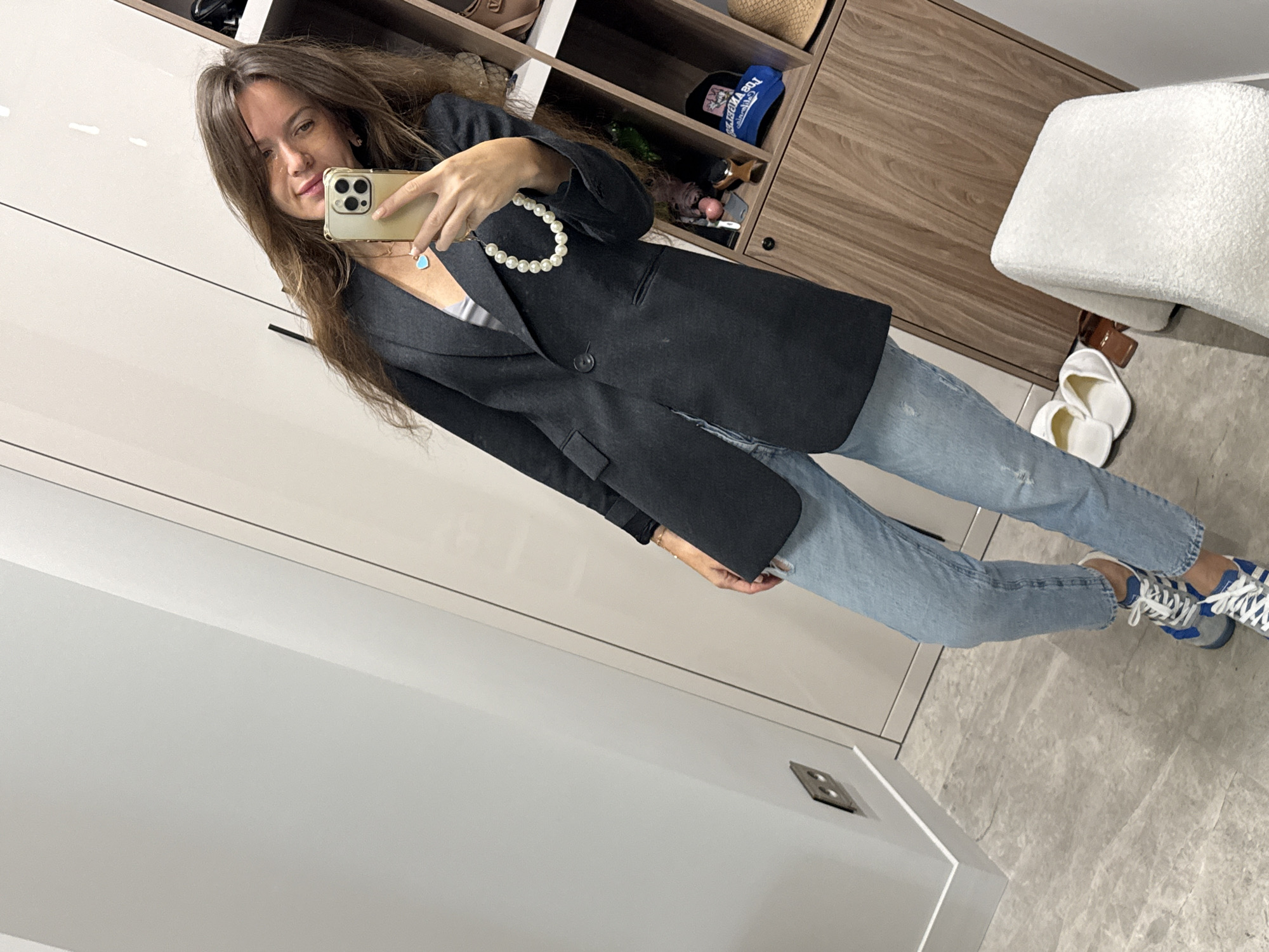 A young woman with long, brown hair is taking a mirror selfie in what appears to be a walk-in closet or dressing room. She is wearing a dark grey blazer over a light grey top and light blue distressed jeans. Around her neck, she has a pearl necklace and a blue pendant necklace. She is holding a gold iPhone in her right hand to take the picture. Her left hand is resting on her blazer. On her feet, she is wearing blue and grey sneakers. To her right, a wooden shelving unit is visible, filled with various items including hats, bags, and other accessories. In the foreground, to her left, are two white slippers and a brown object. A white, textured stool is visible in the lower right corner of the frame. The walls appear to be light colored, with a reflective surface. The overall lighting is bright and even. This scene is located in Moscow, Russia.
