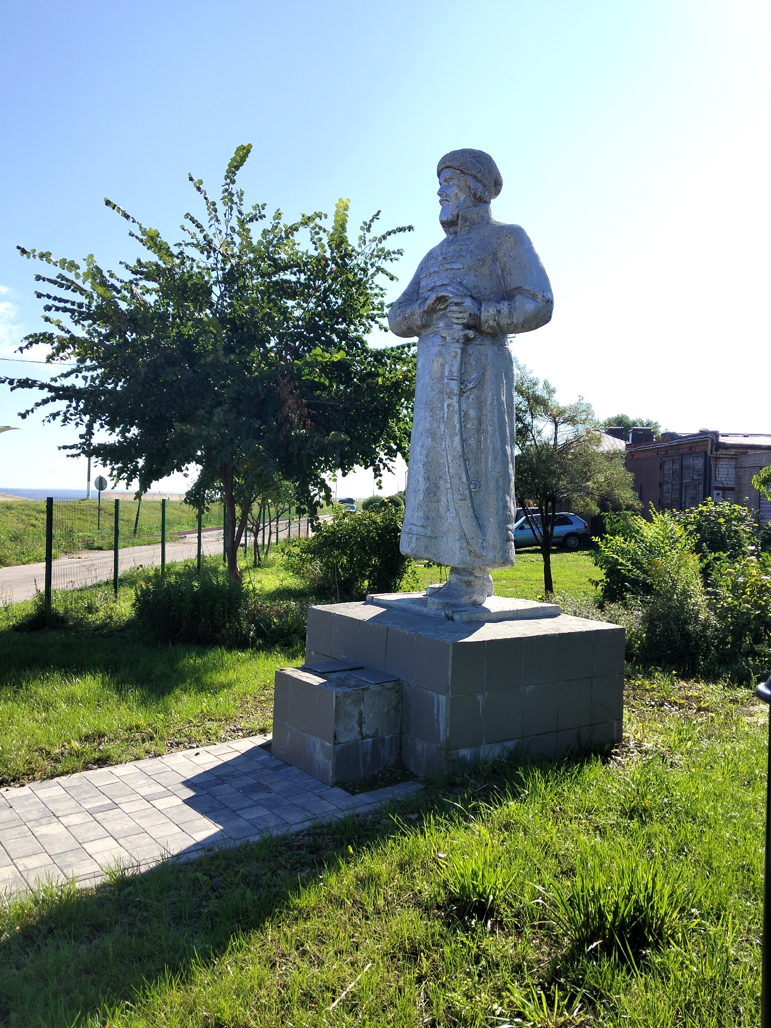 The image shows a statue of a man in historical attire, standing on a multi-tiered concrete pedestal. The statue is made of a silvery-colored material and depicts the man with a beard, wearing a hat and a long coat, with his hands clasped over a sword or dagger. He is facing to the left of the frame.
The statue is situated outdoors in a grassy area with a paved walkway in the foreground. To the left of the statue, there is a green metal fence, a road, and beyond that, a body of water and the sky. Behind the statue, there are trees and bushes, and a brick building is visible in the background. A light blue car is parked on the grass behind some trees.
The scene appears to be a public space, possibly a park or a historical site. The lighting suggests it is a sunny day. The statue is the central subject of the image, with the surrounding environment providing context. The location is identified as being in the city of Kozmodemyansk, Russia.