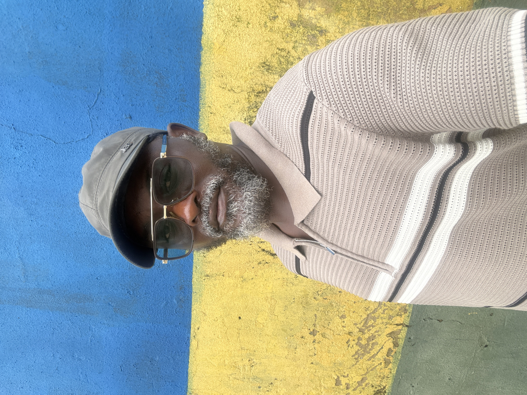 The image shows a close-up of a Black man wearing a grey cap and sunglasses. He has a beard and is looking slightly to the side. He is wearing a light-colored, textured sweater with dark and white stripes on the sleeves and collar. The background is a painted wall with a horizontal stripe of bright blue and yellow. The painting appears to be on an exterior wall. The man is centered in the frame, and the lighting suggests it is daytime, likely sunny given the shadows. The overall mood is casual and relaxed. No visible text is present in the image. The location context provided is Ikoyi, Nigeria.