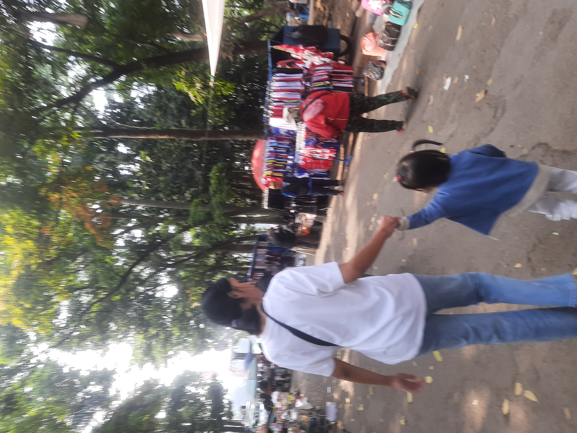 The image depicts an outdoor market scene on a sunny day. In the foreground, a woman wearing a white t-shirt and blue jeans walks while holding the hand of a young child in a blue jacket and white pants. The child's hair is in two pigtails. Behind them, to the left, is a bustling market with various stalls displaying goods. Further back, several people are seen browsing or working at the stalls, and racks of clothing are visible. Trees line the pathway, creating a shaded environment. The ground is paved and scattered with fallen leaves. The overall atmosphere suggests a casual outing at a local market.