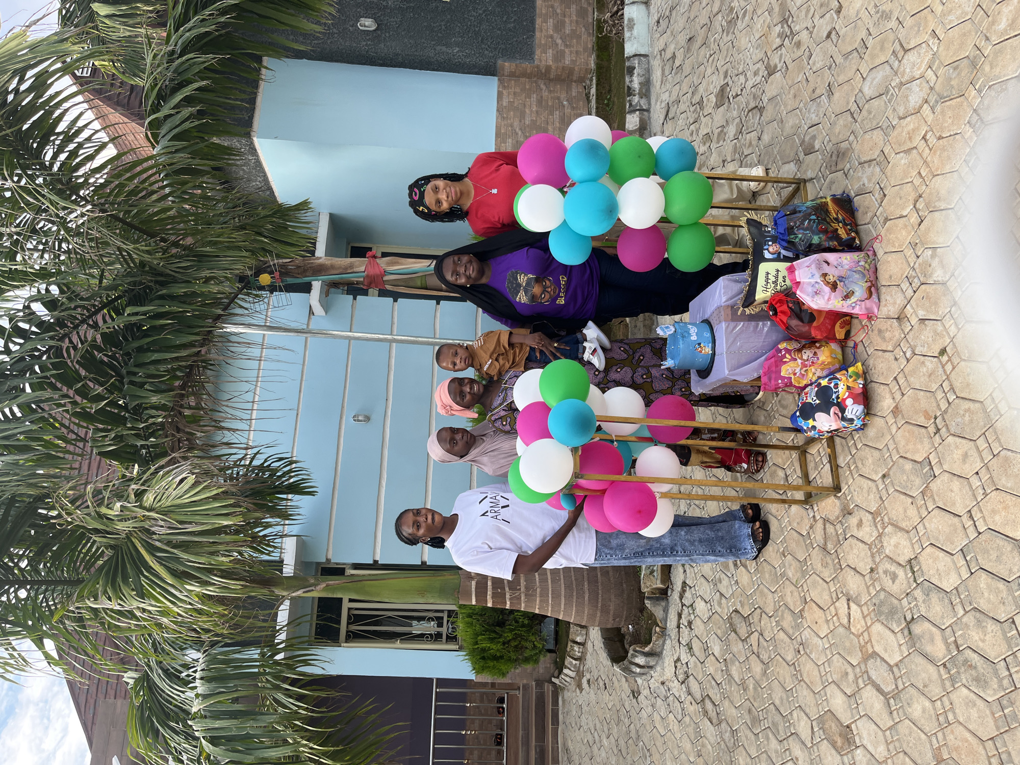 This image captures an outdoor birthday celebration in Jos, Nigeria, featuring six individuals—five females and one male child—gathered on a paved area in front of residential buildings.
On the far left, a female with dark braided hair, wearing a white short-sleeved t-shirt with "ARMANI EXCHANGE" text, distressed blue jeans, and black flip-flops, stands beside a gold-colored frame supporting multiple pink, white, green, and light blue balloons. To her right, a second female, dressed in a light brown hijab and patterned gown, holds a male toddler. The toddler wears a brown t-shirt, blue jeans, and white sneakers, looking towards the left. A third female, adorned in a pink hijab and patterned gown, stands to the right of the second female, smiling towards the camera.
In the central foreground, a small, white-draped table holds a round blue birthday cake with white frosting, a "Happy Birthday" topper, and "Billy" inscribed on its side. Behind the table, a black square pillow displaying "Happy Birthday Son" and a photographic image is visible. On the ground in front of the table are several colorful drawstring gift bags featuring various animated characters, including Disney Princesses, Mickey Mouse, and superhero themes.
To the right of the central display, a fourth female, characterized by dark hair and a colorful headband, wears a purple t-shirt depicting a stylized female figure and the word "BLESSED," paired with dark pants. Adjacent to her, a fifth female, also with dark hair and a colorful headband, wears a red long-sleeved top, light-colored pants, and a necklace with a pendant. Both of these individuals stand near a second gold-colored frame holding an arrangement of white, green, and light blue balloons.
The background consists of light blue painted residential structures featuring brown roofs, white-framed windows, and a section of dark grey textured wall on the right. Several large green palm trees are prominent, with one tree trunk displaying a red ribbon and a small yellow attachment. The ground is uniformly covered with light brown/beige interlocking concrete pavers arranged in a hexagonal pattern. A silver metal railing and green foliage are visible on the left side of the scene, proximate to the building.
