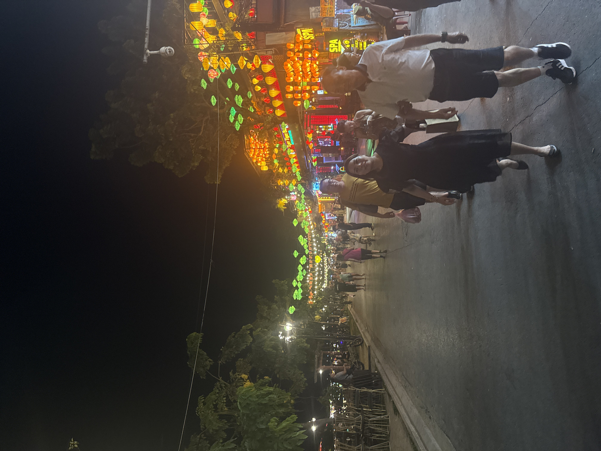This photograph captures a night scene in an urban area, most likely a tourist destination, in an unknown city. A street lined with trees is illuminated by numerous colorful lanterns strung above, leading to a row of commercial buildings, some of which are labeled "SPA" in prominent signage. Pedestrians are seen walking along the street, including a group in the foreground comprised of several people; one wears a white shirt and dark shorts, while another is dressed in a black dress. The environment is dark save for the street and surrounding buildings which are brightly lit up.