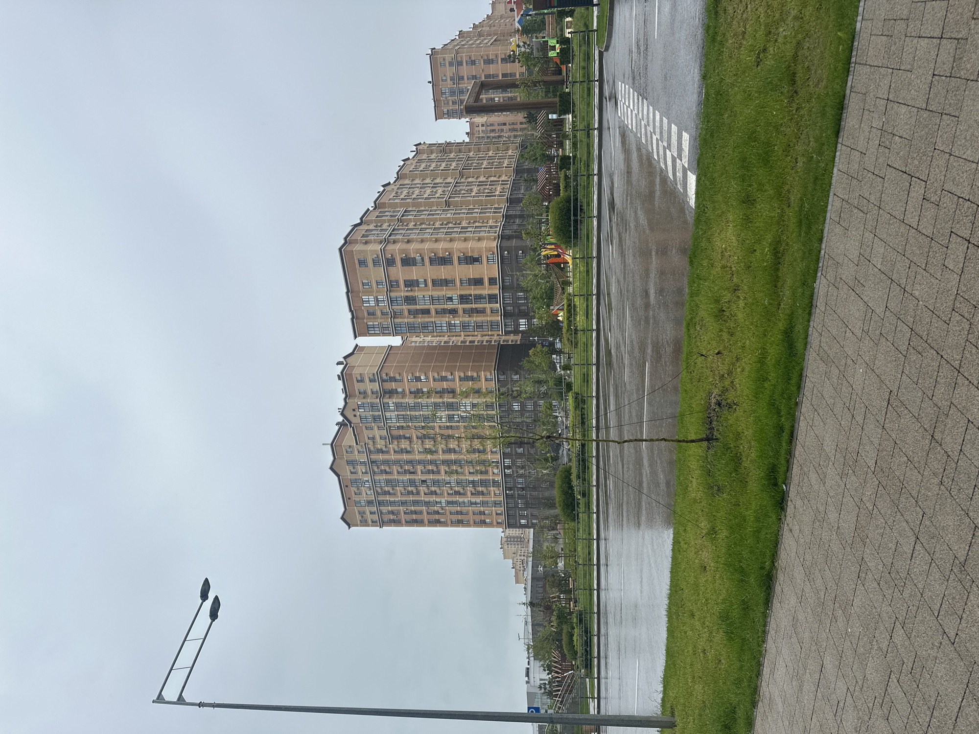 The image depicts a wet, overcast day in Stavropol, Russia. Tall, tan-colored apartment buildings line a street with marked parking spaces. Between the parking area and a brick sidewalk, there is a strip of green grass. A bare sapling tree stands in the grassy area. A street lamp is visible in the foreground. No people are visible in the scene.