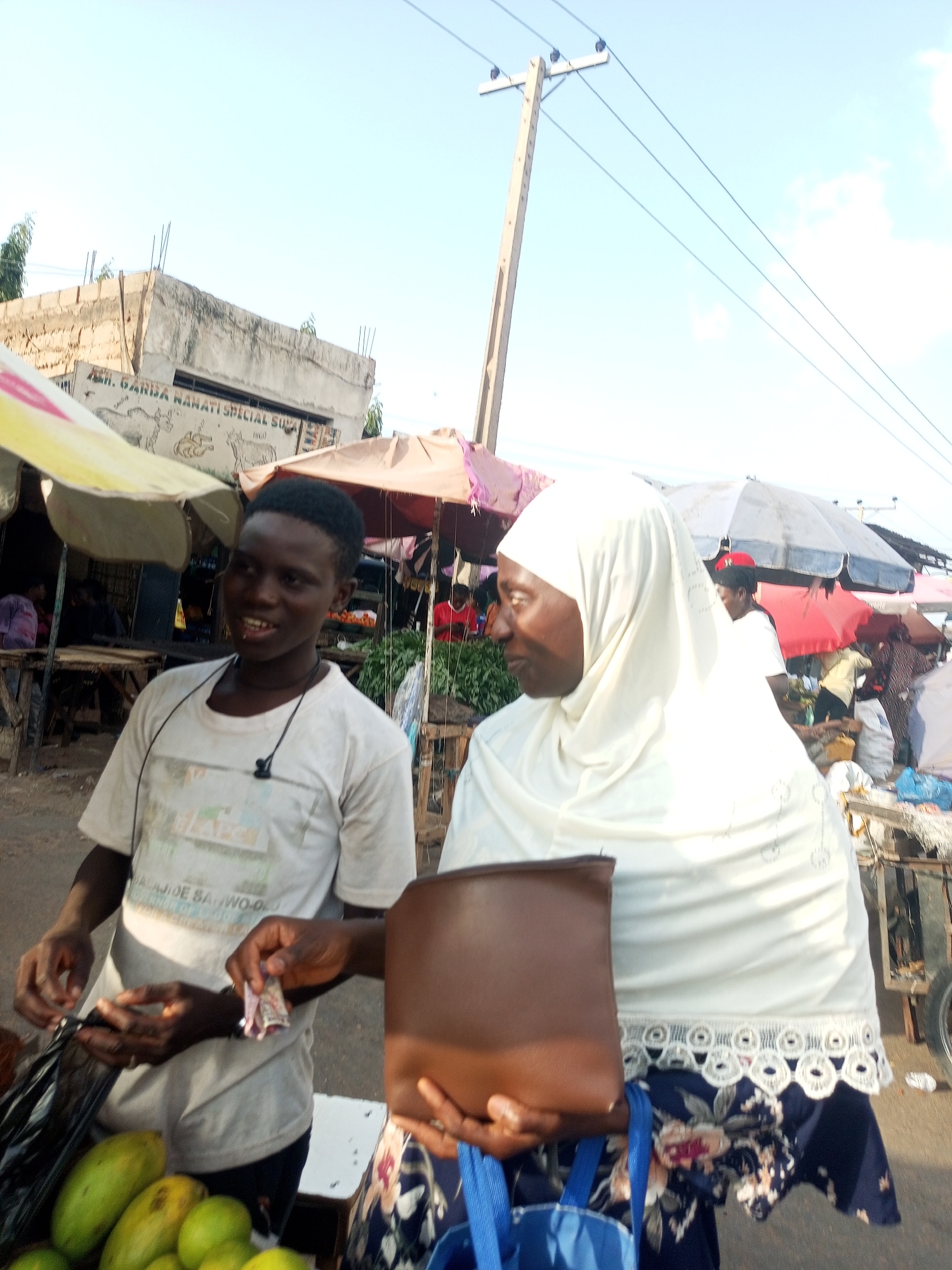 In an outdoor market scene, a young Black male, appearing to be a vendor, engages with an older Black female customer. The vendor, wearing a light-colored t-shirt with a graphic print and a black cord around his neck, smiles and holds out a small wad of cash in his left hand. His right hand is near a black plastic bag, possibly preparing to bag purchased items. In front of him are several green fruits, likely mangoes. The customer, draped in a white hijab and a white garment, faces the vendor and holds a brown purse in her right hand and a blue bag with her left. She appears to be looking towards the vendor or at the transaction.
Behind them, the market bustles with activity. Colorful umbrellas provide shade over stalls laden with produce, including leafy green vegetables. Several other individuals are visible in the background, some tending to stalls or walking through the market. A building with visible construction elements and signage is situated in the mid-ground, and a utility pole with wires stretches across the sky.
The lighting suggests it is daytime, likely late afternoon due to the warm tones and long shadows. The sky is bright with a few scattered clouds, indicating fair weather. The overall atmosphere appears to be one of a typical marketplace interaction, with the focus on the exchange between the vendor and customer.
Visible text on the building in the background reads: "MR. GARBA NAMATI SPECIAL SOU". The t-shirt worn by the vendor has some illegible text or graphic.
