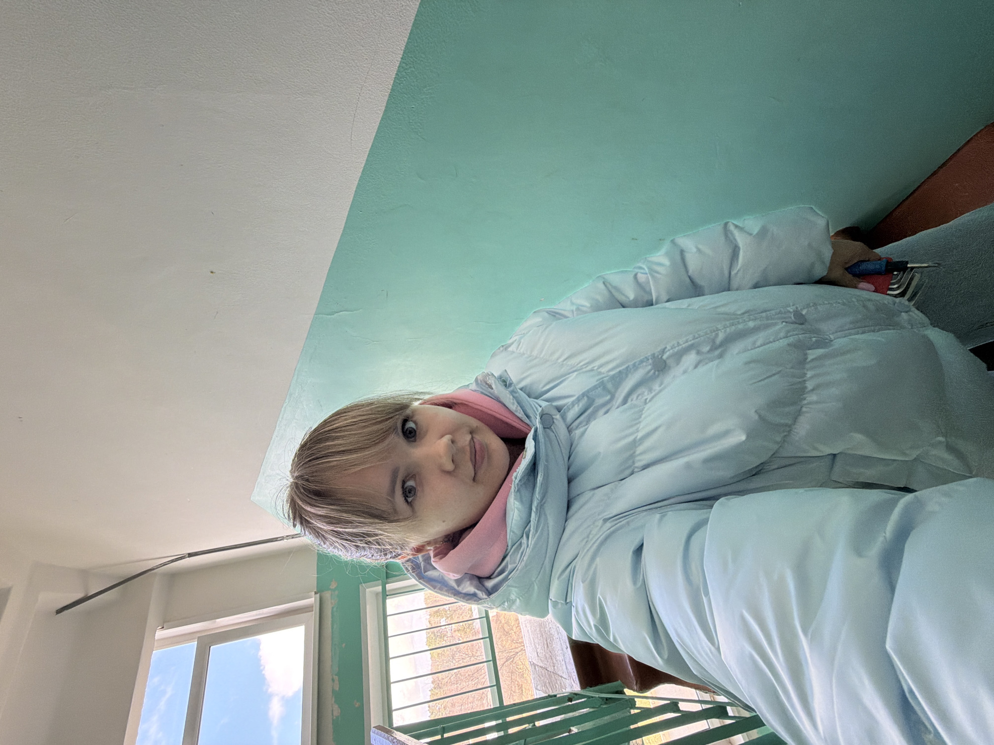 The image is a selfie taken indoors, possibly in a stairwell of a building in an unknown city. A young woman is looking into the camera, wearing a light blue puffer jacket over a pink hooded sweatshirt. She is holding a small, red and blue tool, possibly a screwdriver or similar hand tool. The walls are painted in two tones: white on the upper portion and a light greenish-blue below. A window is visible in the background, offering a view of the sky and some greenery, suggesting an exterior setting. The woman appears to be standing on or near metal stairs with a green railing.
