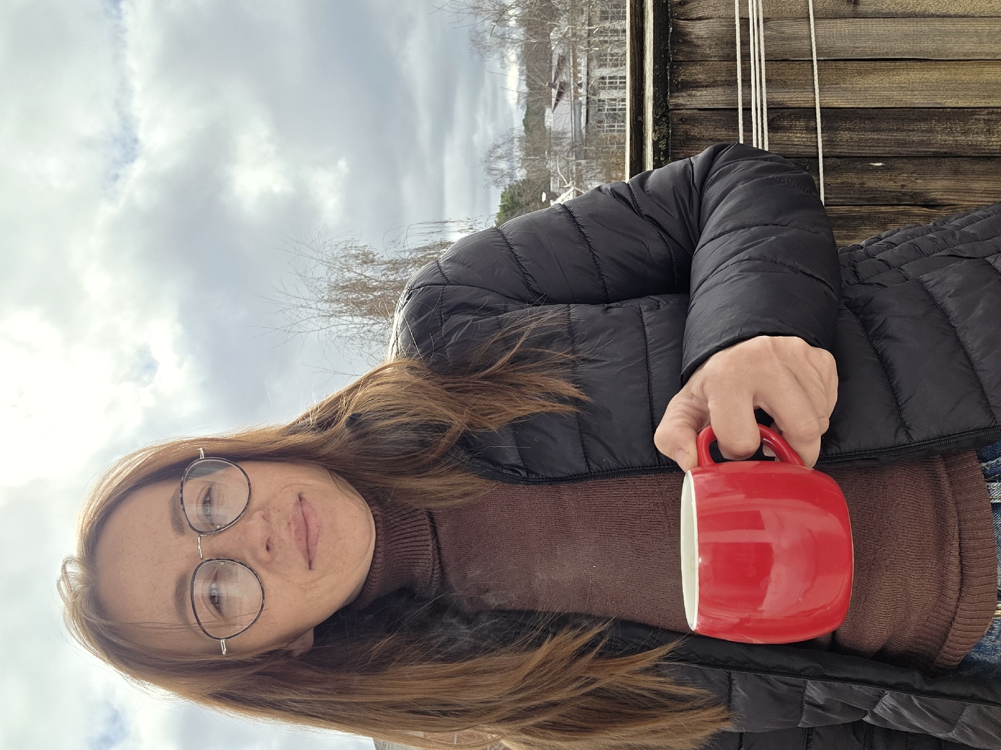 A woman with long brown hair, wearing glasses, a brown turtleneck sweater, and a black puffer jacket, is holding a red mug. She is standing outside near a wooden fence. In the background is a cloudy sky and a building covered in snow. This location appears to be in the city of Chaikovsky, Russia.