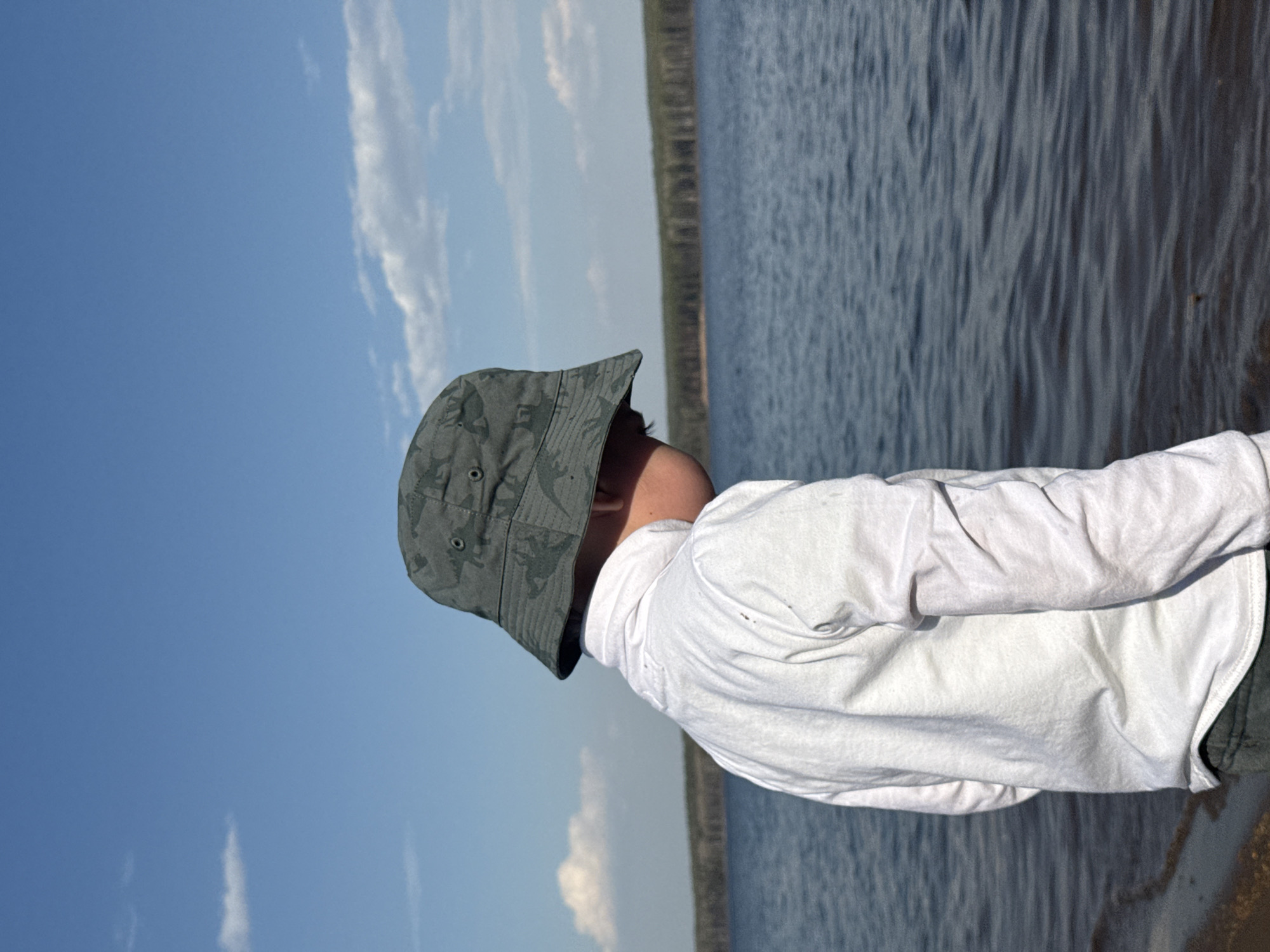 A person wearing a camouflage bucket hat and a long-sleeved white shirt stands near a body of water. In the background are trees and a blue sky with light clouds. Based on the question, this location is in the city of Chaikovsky, Russia.
