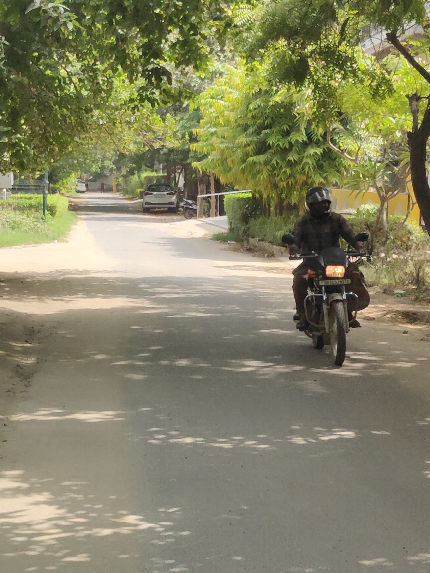A man wearing a helmet and a dark patterned shirt rides a motorcycle down a tree-lined street in Gurgaon, India. The street is paved and appears to be an asphalt road. On the left side of the road, there is a grassy verge with some bushes and trees. On the right side of the road, there are more trees and some buildings in the background. The motorcycle has its headlight on and a license plate that reads "HR 26 DJ 6926". The scene is brightly lit by sunlight, which casts shadows on the road from the trees overhead. In the background, a white car is parked on the side of the road, and a black scooter is also visible. The overall environment suggests a suburban or residential area within the city.