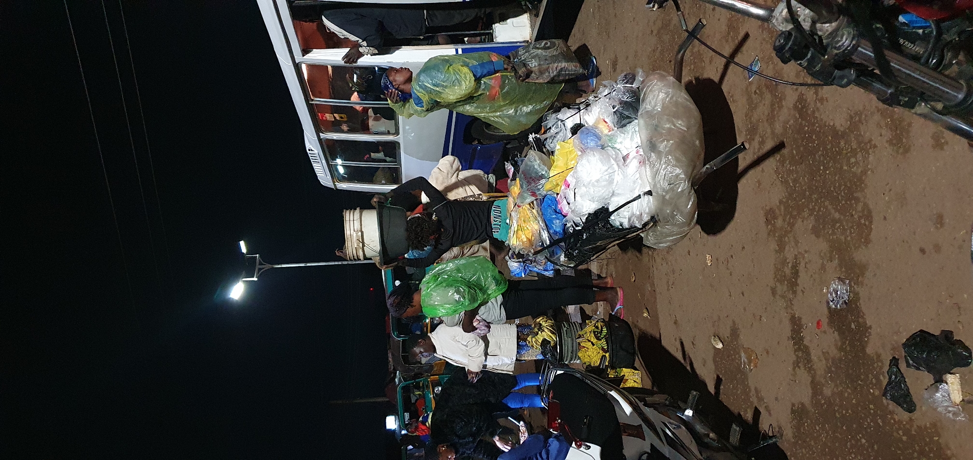 In Bukuru, Nigeria, at night, under a street lamp, several people are gathered around a bus. A woman is balancing a large black basin on her head, filled with items, and is wearing a black long-sleeved shirt with "NON-STOP" visible on it. She also has a green plastic bag over her shoulder. Another woman beside her wears a light-colored jacket and is looking away. A person in a yellow plastic poncho stands near the bus door, holding a dark bag. A wheelbarrow filled with plastic-wrapped items, including what appears to be textiles in yellow, blue, and white, is positioned in the foreground. A white bus is parked, with its door open and passengers visible inside. In the background, several green and white "Keke Napep" (auto-rickshaws) are parked. The ground is muddy and littered with debris. A motorcycle is parked to the left, and another motorcycle's handlebars are visible in the lower right corner.
