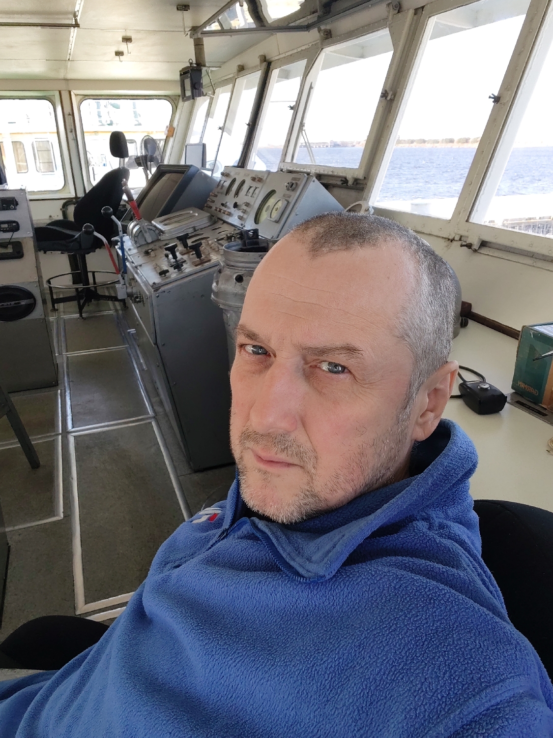 The image captures a selfie of a man seated in what appears to be the bridge or pilothouse of a vessel. The subject is a middle-aged man with short, thinning grey hair and a short beard or stubble, looking directly at the camera with a neutral expression. He is wearing a blue fleece jacket with a small, indistinct red and white logo or patch on the left chest.
The setting is the control room of a ship or boat, characterized by a complex navigation console in the background. This console features numerous dials, gauges, switches, and levers, as well as a steering mechanism. A tall chair is visible behind the console on the left. Large windows surround the bridge, providing ample natural light and a clear view of the exterior.
Outside the windows, calm water is visible, extending towards a distant shoreline dotted with trees, suggesting a maritime location like a river, lake, or coastal area. The bright light indicates it is daytime with clear weather conditions. On a surface to the right, a black electronic device and a green box with partially visible, possibly Cyrillic, text "НМКАН" are discernible. The man is likely taking a photo of himself in his workplace or during a journey.