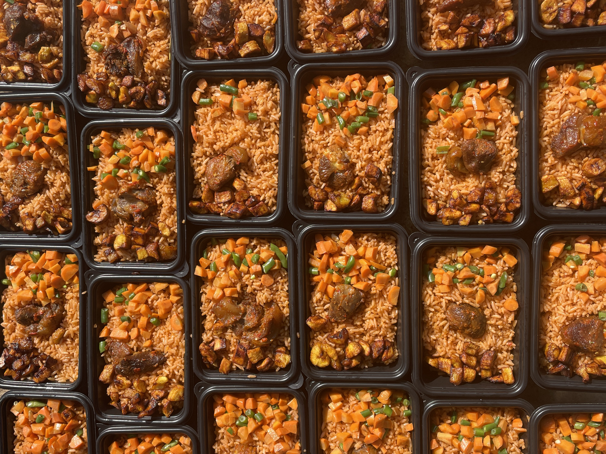 The image displays a top-down, overhead view of multiple black plastic food containers filled with Jollof rice, chopped carrots, and green beans. Some containers also include pieces of meat and fried plantains. The food appears to be prepared and portioned for takeout or meal prep. The scene is not identifiable as being in Jos, Nigeria, as there are no specific geographical markers present. The lighting suggests an indoor or shaded outdoor environment.