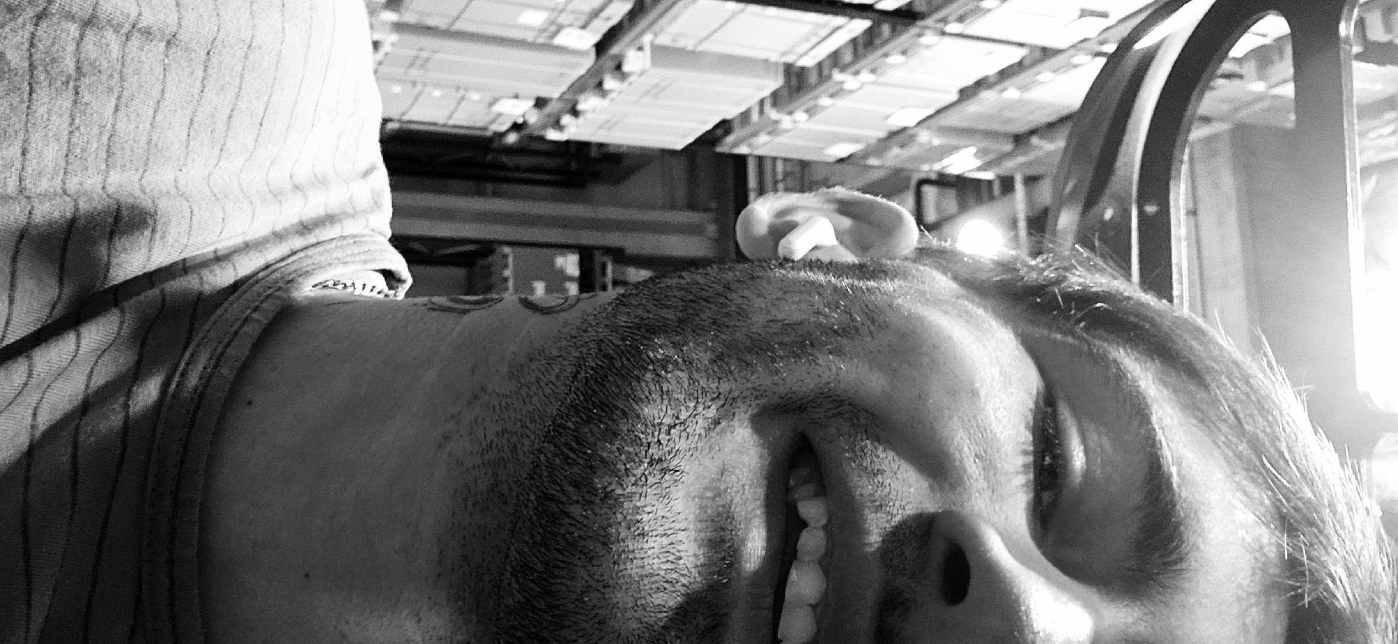 A black and white close-up image shows a man lying on his back, his face turned to the side. The man has a beard and open mouth. His ear is visible near the top of the frame. He is wearing a tank top. In the background, there is a ceiling structure with rectangular panels. There is an open metal doorway. The location is not discernible based on visual information alone.