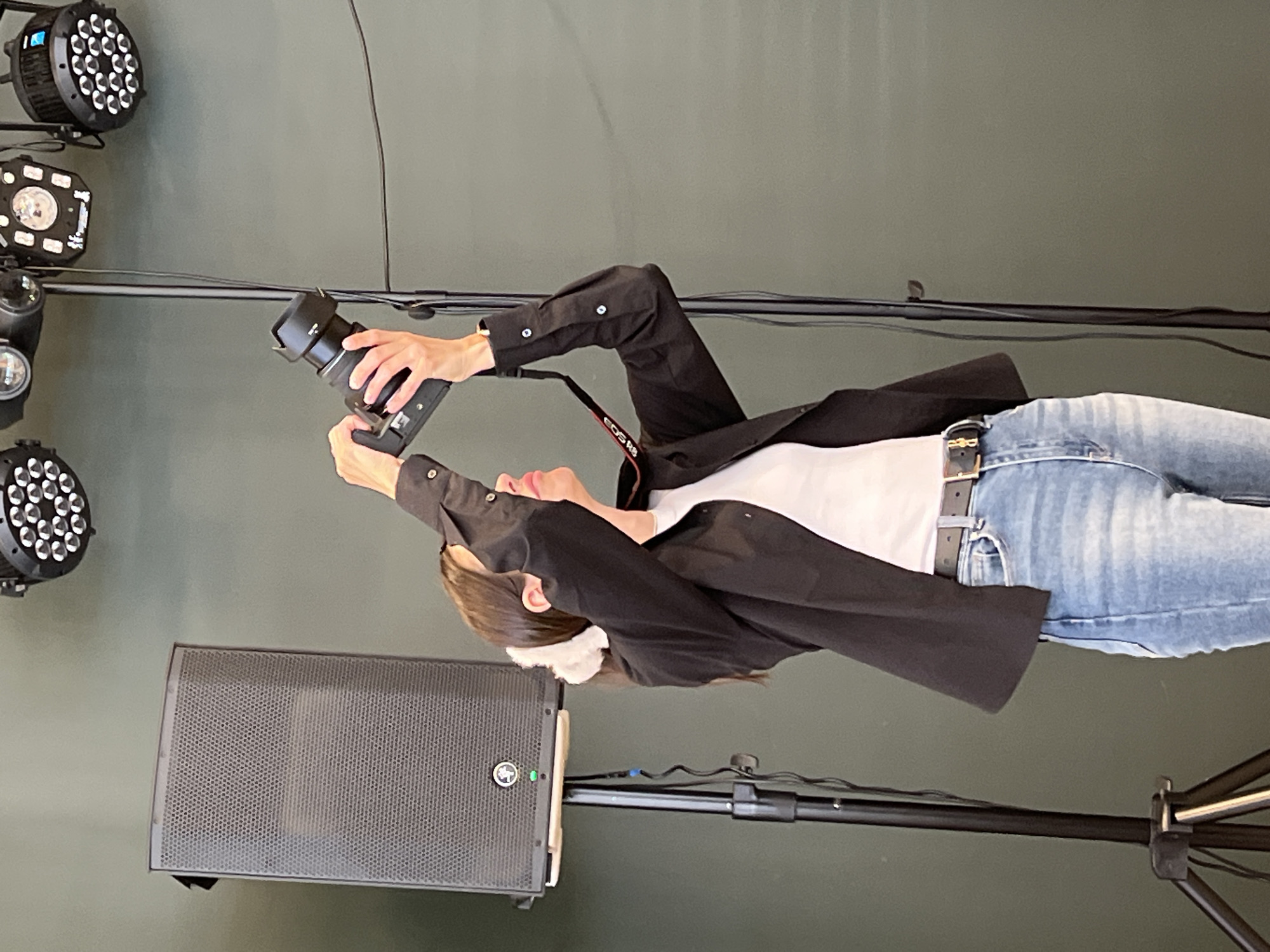 A woman is holding a camera and looking up. The woman is wearing a black blazer, a white t-shirt, and blue jeans. The location appears to be indoors, possibly a studio or event space, indicated by the lighting equipment and speaker visible in the image. There is no evidence to suggest that the photograph was taken in Saint Petersburg, Russia.