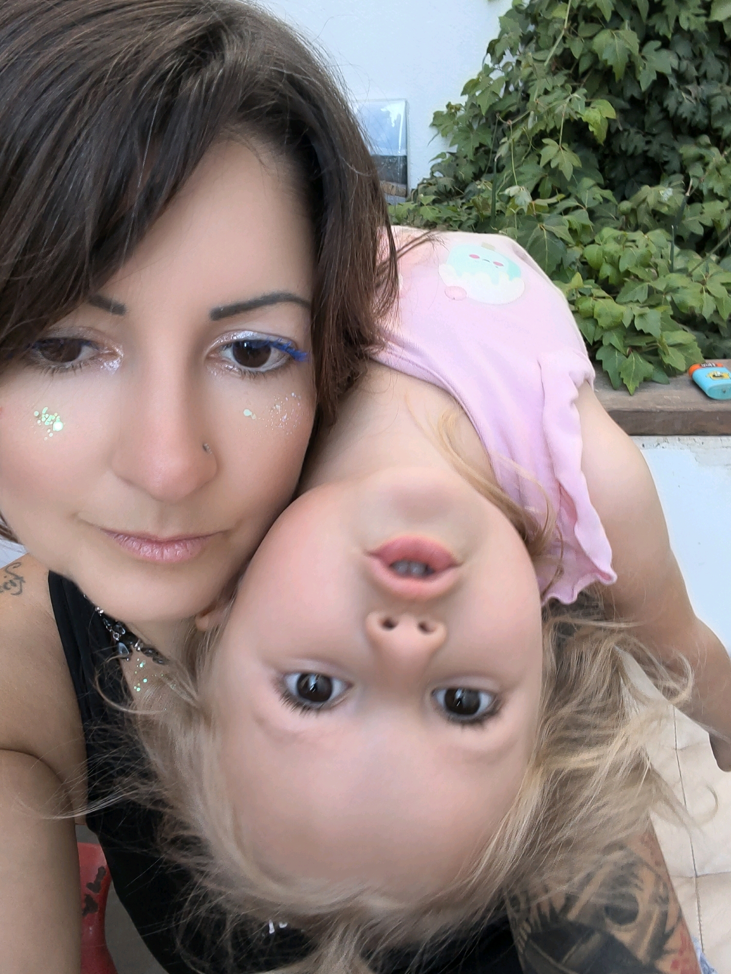 In a close-up, vertical, selfie-style photograph taken outdoors in Lachish Regional Council, Israel, a woman and a young child are depicted. The woman, with dark brown hair and striking blue eyeliner, is positioned on the left, her face tilted downwards. She is wearing a black tank top adorned with a necklace featuring a star charm and what appears to be glitter. Scattered on her face are light green sparkles. The child, whose head is upside down and closer to the camera, has blonde hair and is wearing a pink t-shirt with a cartoon ice cream cone graphic. The background consists of lush green foliage, suggesting a garden or natural setting, and a white wall. A small, blue object, possibly a toy or a dispenser, rests on a ledge to the right. The lighting suggests it is daytime.