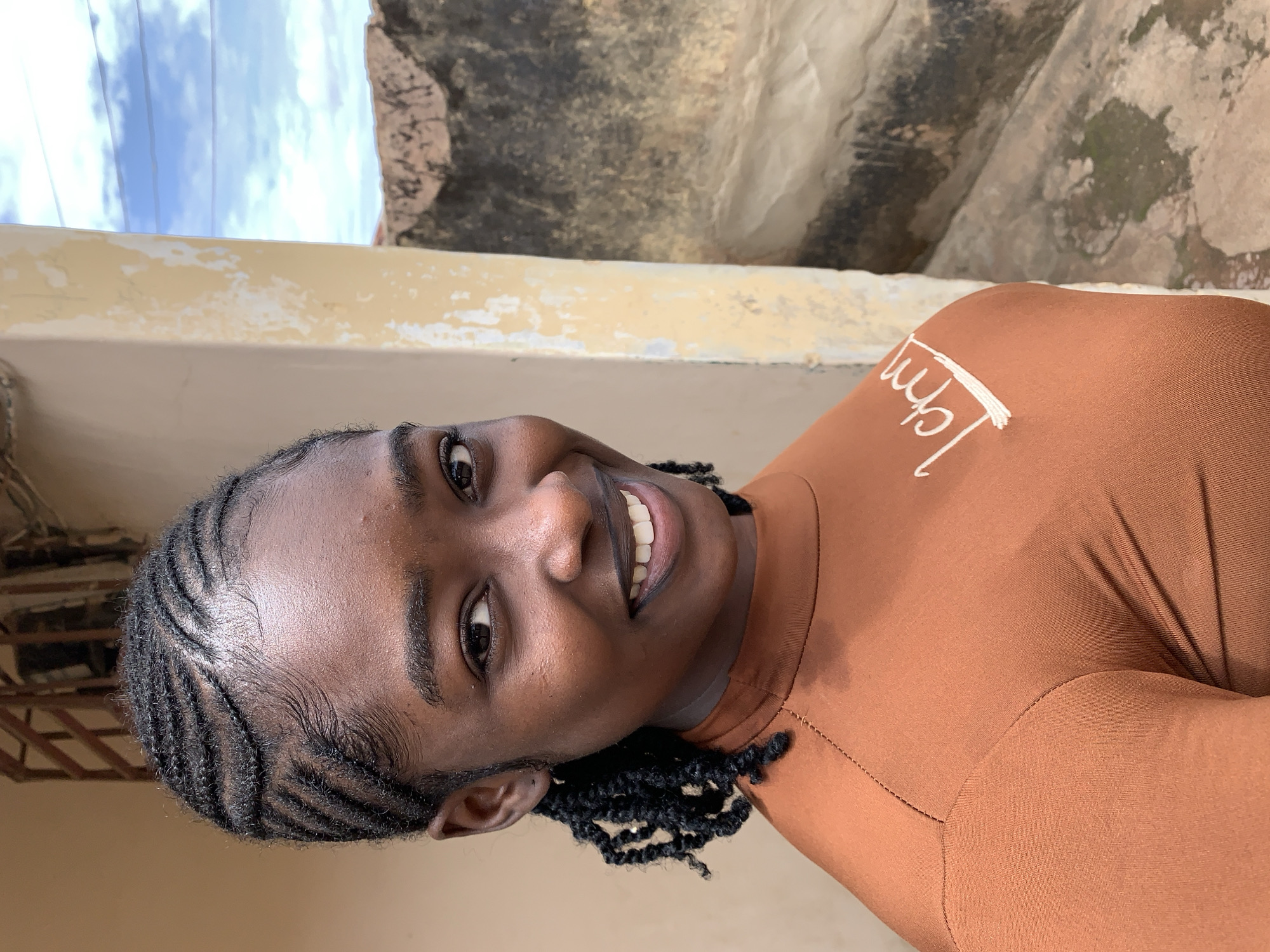A young Black female is depicted in a head-and-shoulders portrait, angled slightly to her right, looking directly at the viewer. Her hair is styled in tight cornrows across the scalp, featuring sculpted baby hairs along the hairline, and extends into twisted braids framing her face and ears. She has dark eyes, full eyebrows, and a wide smile revealing white teeth. Her lips are defined with a dark pigment. She wears a fitted, long-sleeved top of a rust-brown hue, with a high neck and visible stitching along the shoulders and arm seams. White embroidery, appearing as stylized text, is present on the left side of her chest.
The setting is outdoors in Rafin Par, Nigeria. To the subject's immediate left, a plain, light beige wall is visible. To her right, a heavily weathered, light yellow pillar or wall section shows significant paint loss and exposure of a darker, textured underlayer. Beyond this pillar, a bright blue sky with scattered white clouds is visible at the top. Below the sky, a rough, dark-colored, and weathered wall or structure appears in the mid-ground, exhibiting textural irregularities and discoloration. Indistinct ground elements are visible at the bottom right of the frame.