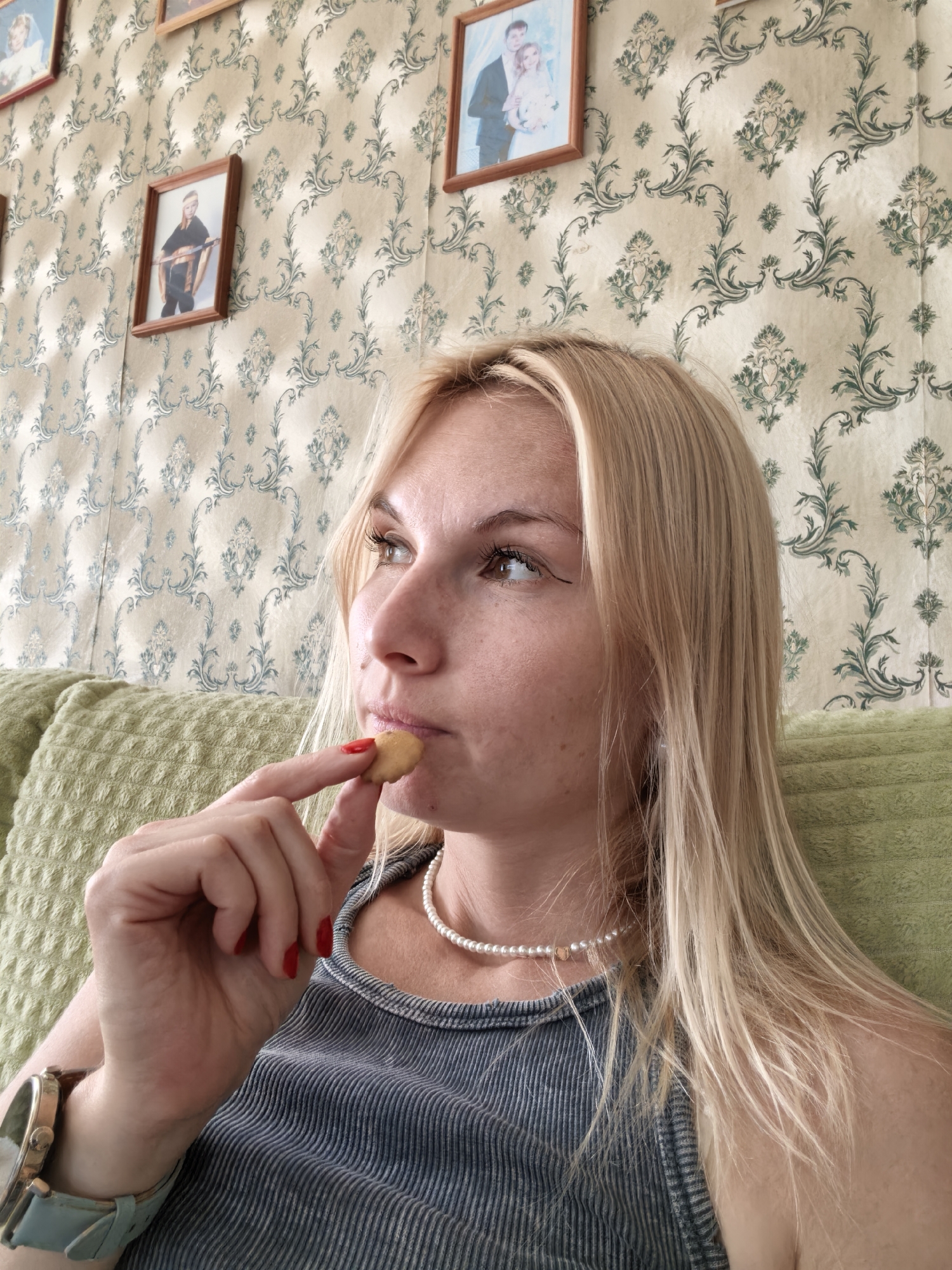 A woman with blonde hair is sitting on a couch in front of a patterned wallpaper. She is holding a small cookie up to her mouth, about to eat it. She is wearing a gray ribbed tank top and a pearl necklace. A watch with a light blue band is visible on her left wrist. In the background, several framed photographs are hung on the wall, depicting people in formal attire. The couch she is sitting on is covered with a textured green fabric. The scene appears to be indoors, possibly in a residential setting. There is no identifiable information linking this image to the city of Sterlitamak, Russia.