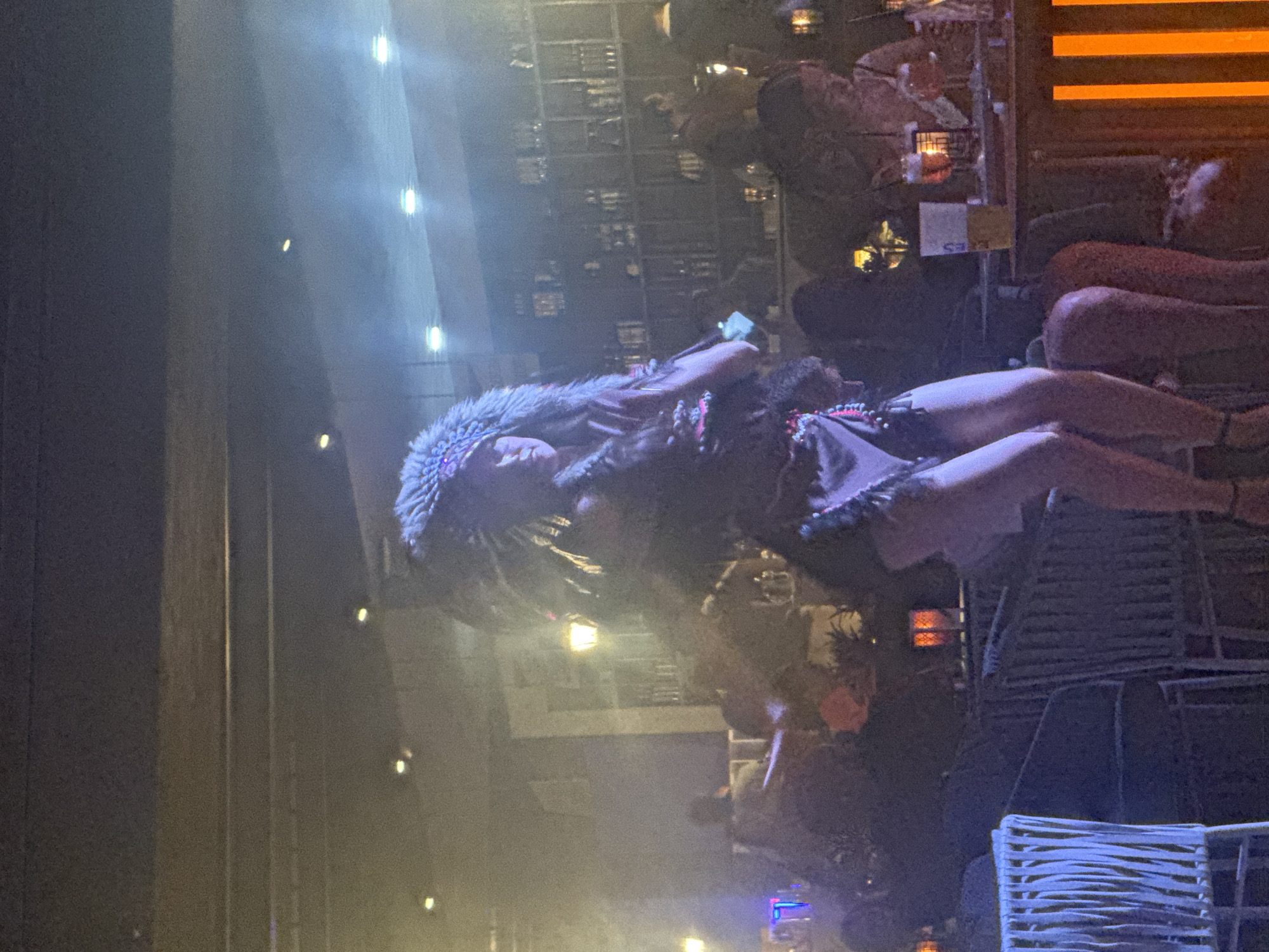 The image shows a performer wearing a headdress and costume, possibly a Native American-inspired outfit, on a stage in a dimly lit venue. The performer is in mid-pose or dance, with their legs visible. In the background, patrons are seated at tables in what appears to be a bar or restaurant. There are bottles and glasses on a bar in the background. The venue has a ceiling with visible lights and shelves filled with bottles. The overall atmosphere suggests a lively entertainment setting. This scene could be from a venue located in the city of Manavgat, Turkey, known for its tourist resorts and entertainment options.