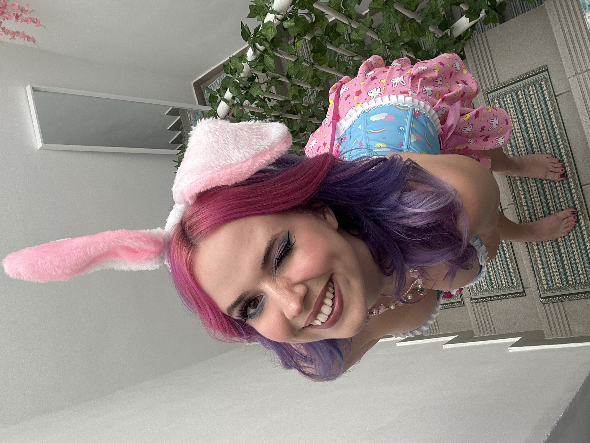 A woman with pink and purple hair, wearing bunny ears and a pink dress with unicorns and moons on it, is smiling and winking at the camera. She is standing on a rug on a set of stairs, with plants in the background. The location is in an interior. The city of Zagreb, Croatia, is not discernible from the image.