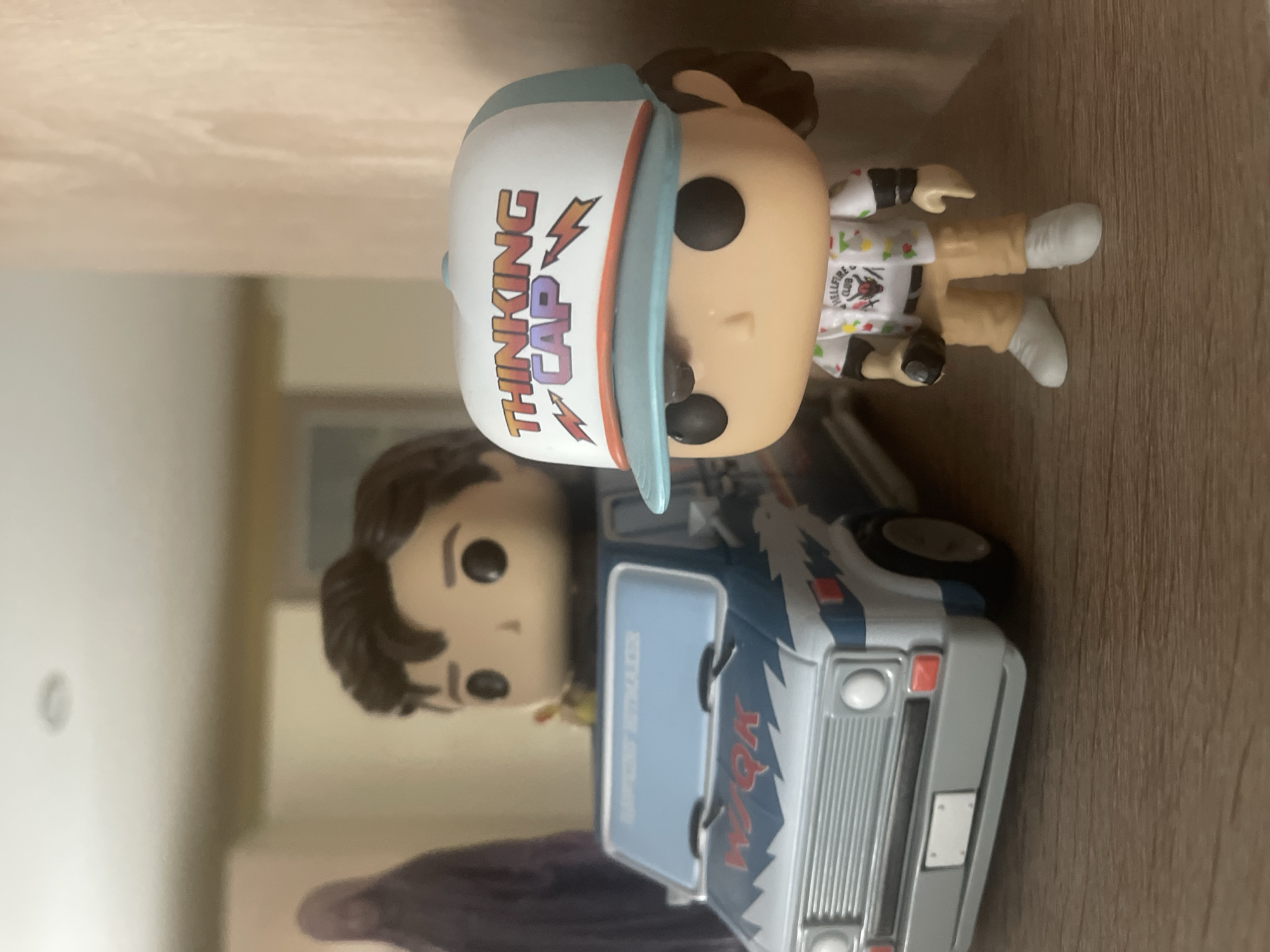 A close-up, eye-level shot shows two Funko Pop figures on a wooden shelf, with a blurry background. In the foreground, a figure with brown hair wears a white trucker hat with blue trim that says "THINKING CAP" in colorful letters, and a white shirt with colorful graphics, including a red demon head. The figure holds a black object in its right hand and wears tan pants and white sneakers. Behind it, another Funko Pop figure with brown hair is seated in a blue and white van with red lightning bolt decals and "WSQK" printed on the hood. The van has a white license plate and a blue windshield with text that appears to be "RAPTOR RETRIEVER". The background is out of focus, showing a wall and a faint image of a person in a dark hooded robe. The overall lighting is soft and warm, coming from an unseen source. This scene is not located in the city of Khimki, Russia.