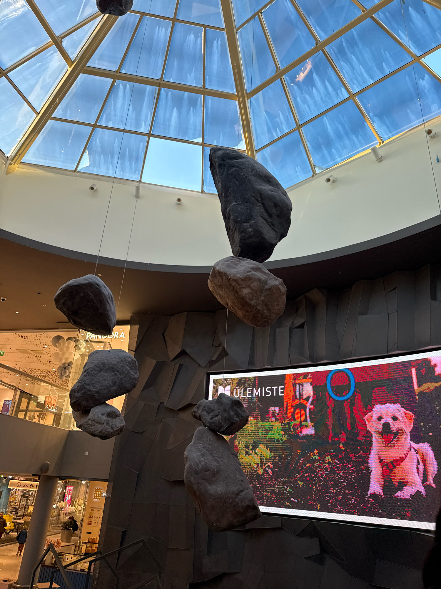 The image shows an interior view of a shopping mall with a modern architectural design. In the foreground, several large, grey rocks are suspended from the ceiling by wires, appearing to float in the air. Behind the rocks, a large digital screen displays an image of a white dog and text that reads "ÜLEMISTE". The screen is mounted on a dark, angular wall. Above the screen and rocks, a large, geometric skylight composed of glass panels and metallic framing allows natural light to enter the space. In the background, visible through openings and glass facades, are various shops and escalators, indicating a bustling commercial environment. The mall appears to be Ülemiste Keskus, located in Tallinn, Estonia, as suggested by the text on the screen. The scene is well-lit, with a mix of natural and artificial light sources.