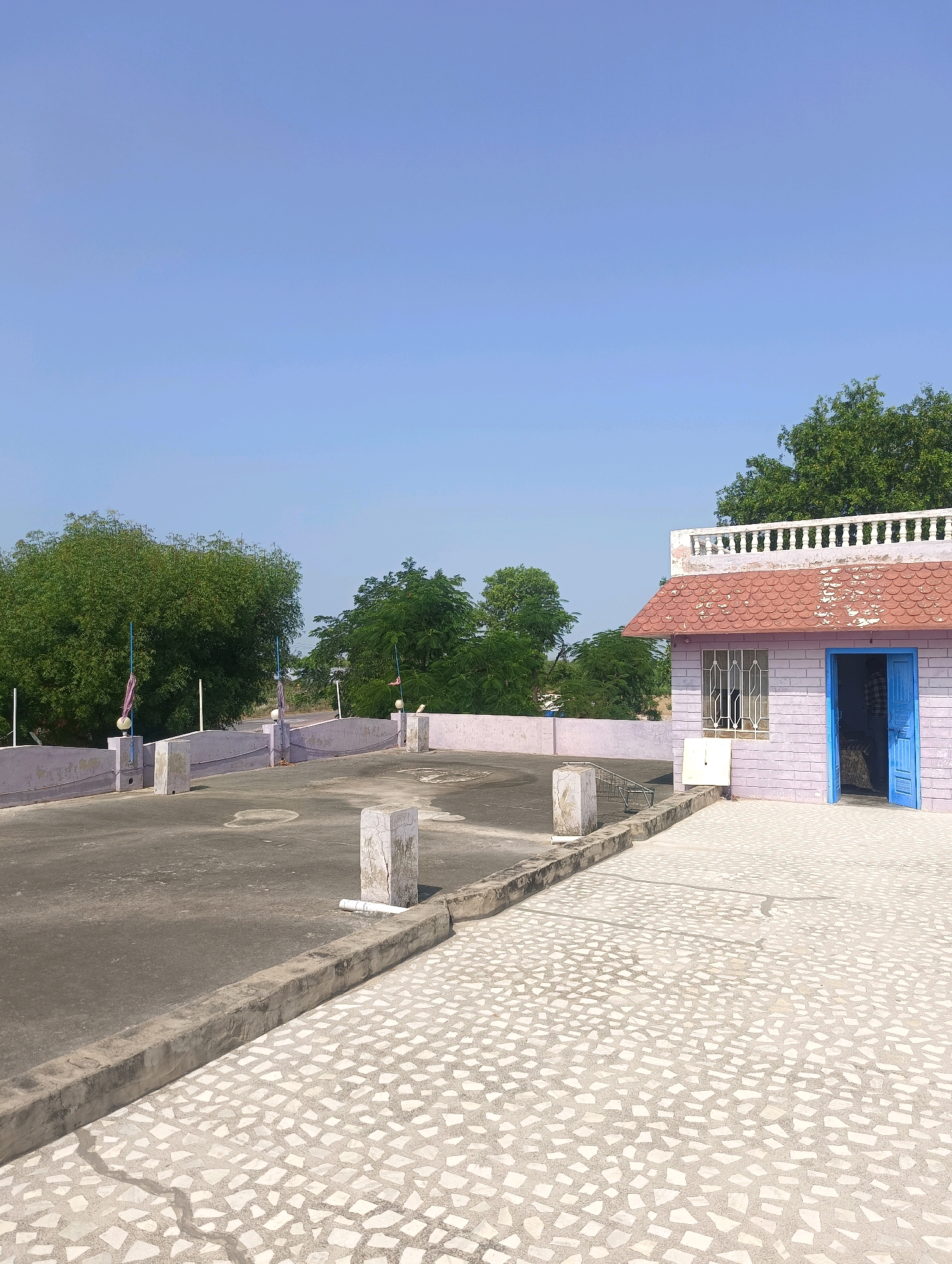 This is a photograph of a rooftop in Chhapri Bari, India. The rooftop is divided into two sections: a darker, paved area in the foreground and a lighter, tiled area in the background. A small, lavender-colored building with a red tiled roof is situated on the right side of the image. The building has a blue door that is open, revealing a person inside. There are trees and a clear blue sky in the background. Several concrete pillars and poles are visible on the rooftop, some with flags attached. The scene suggests a warm, sunny day.