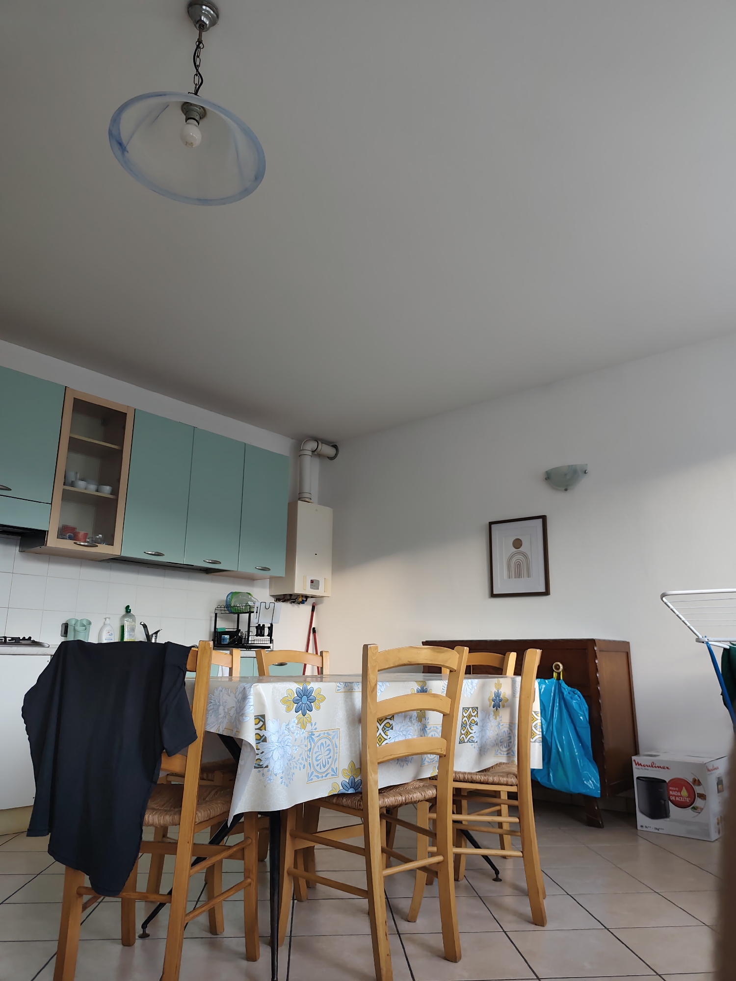 This is an indoor shot of a dining area and kitchen in what appears to be a residential setting, possibly in Guarene, Italy. The scene is illuminated by natural light, suggesting it is daytime.
The main subjects are a wooden dining table covered with a floral patterned tablecloth and four wooden chairs. A black t-shirt is draped over one of the chairs. The table is set for a meal, with plates and possibly cutlery partially visible.
In the background, a pale blue kitchen cabinet unit is mounted on the wall, featuring a glass-fronted section showcasing white ceramic dishes. Below the cabinets is a white tiled backsplash with a sink and various cleaning supplies. A gas water heater is mounted on the wall to the right of the cabinets.
Further to the right, a dark wooden dresser stands against the wall. On top of the dresser is a framed piece of art with a minimalist rainbow motif. Next to the dresser, a bright blue trash bag is tied up and placed on the floor. To the far right, a box labeled "Moulinex" is visible, along with part of a white clothes drying rack.
The ceiling has a single pendant light fixture with a light blue frosted glass shade. A wall-mounted light fixture is also visible above the dresser. The floor is tiled with a beige grid pattern. The overall impression is of a functional and lived-in space.