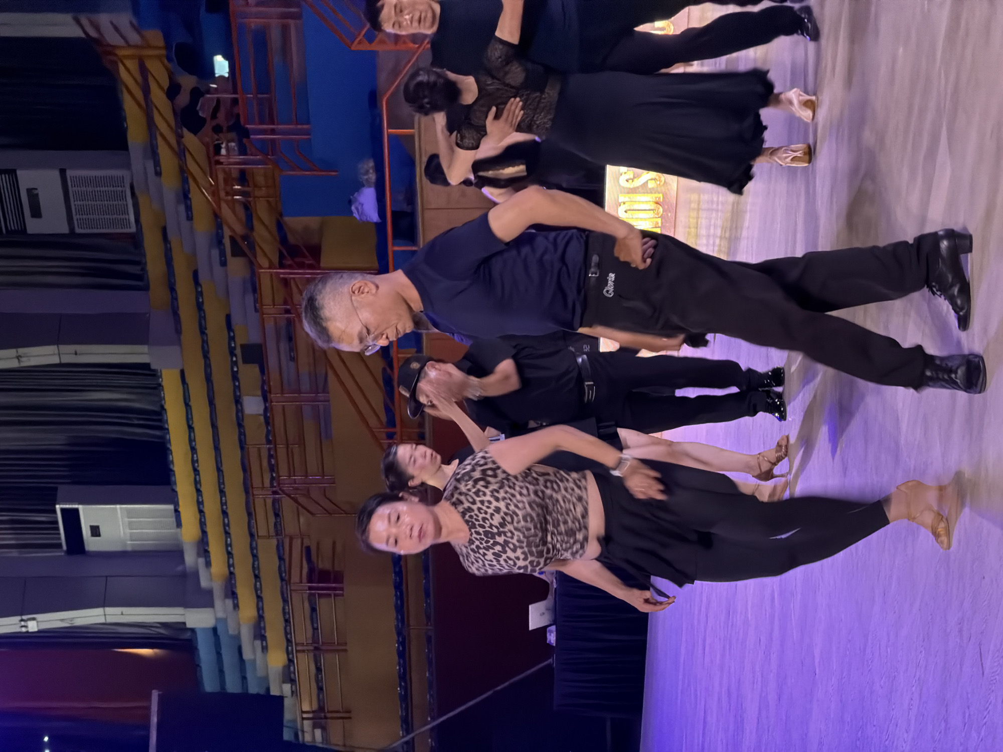 In a ballroom, individuals are engaged in dancing. A woman in a leopard-print top and black skirt is prominently positioned in the foreground, with a man in a dark blue shirt and black pants to her right. Other dancers, in black attire, are visible in the background. The environment features tiered seating with yellow and blue accents, a large air conditioning unit, and a stage with a backdrop of curtains. The floor is illuminated with purple lighting. The scene appears to be an indoor dance competition or performance.