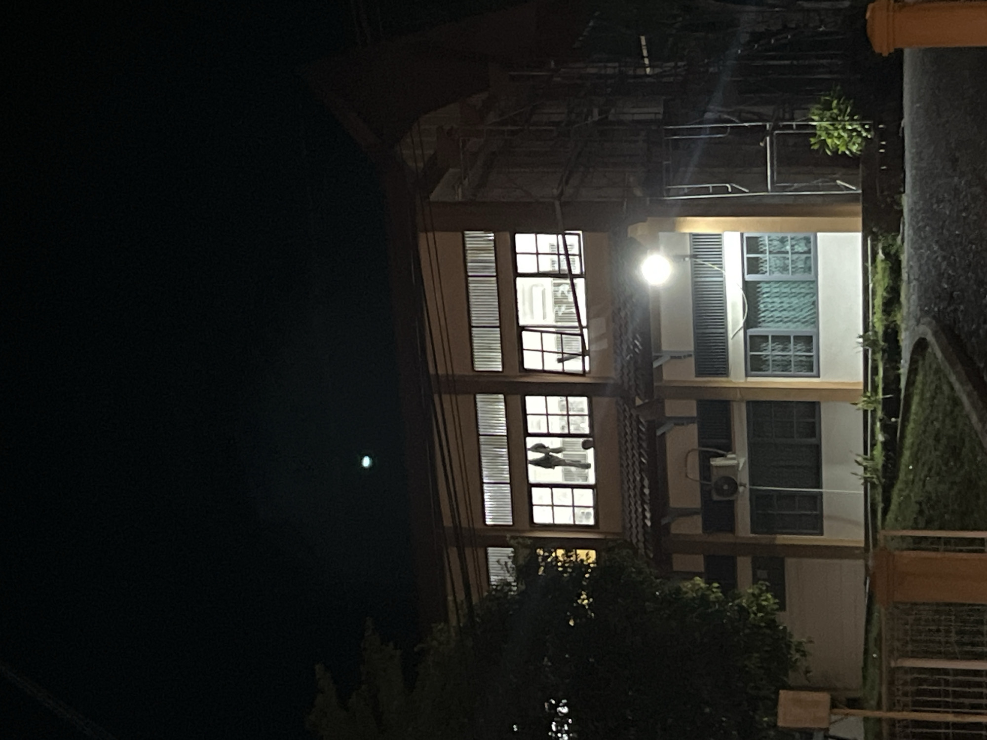 The image shows a building at night in Lhokseumawe, Indonesia. The building has multiple stories with windows, some of which are illuminated from within. A streetlight is visible near the building, casting light on the structure and nearby vegetation. There are trees and greenery in the foreground. The sky is dark.