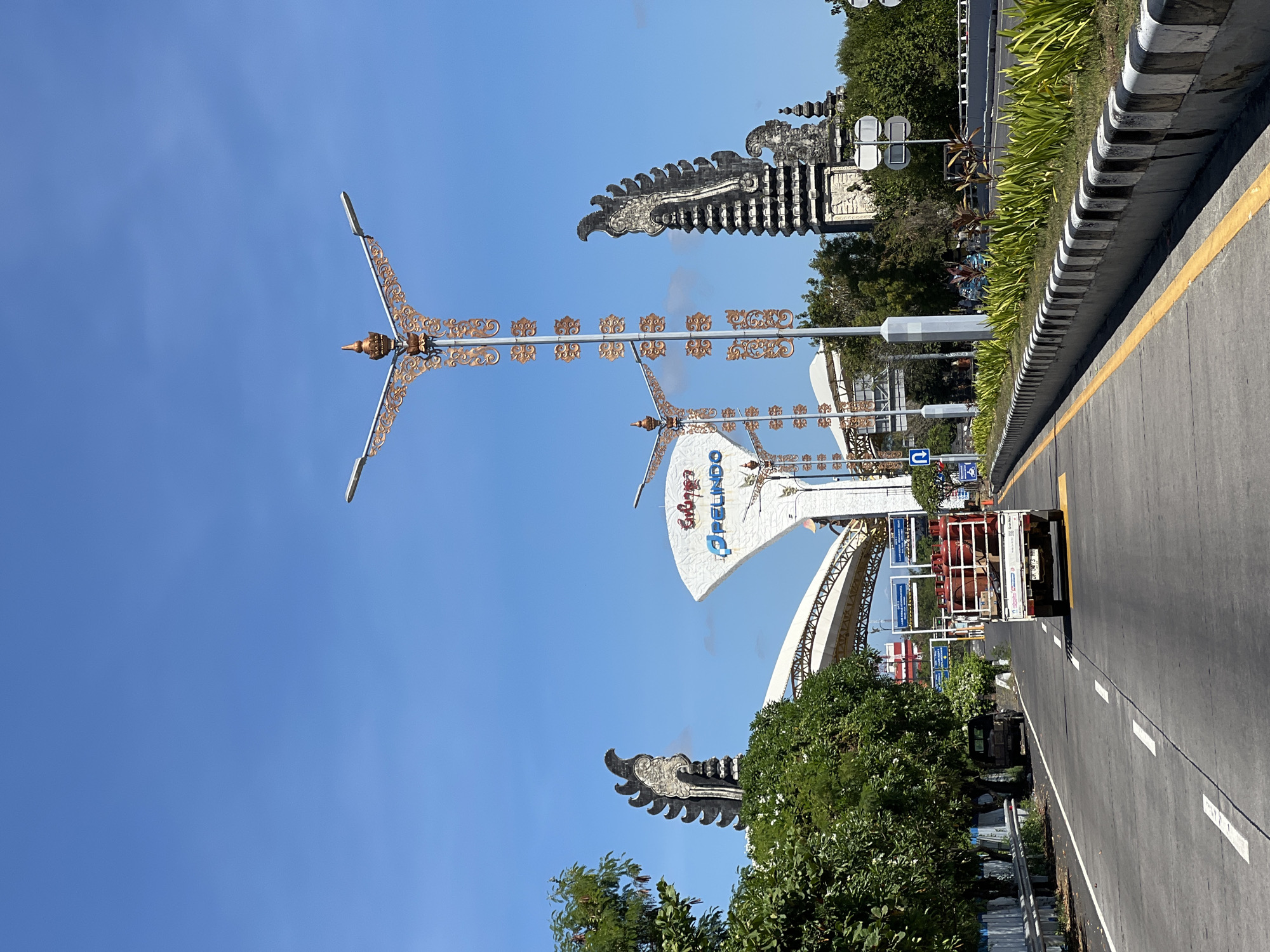 An eye-level, outdoor, vertical, medium shot captures a bright, sunny day on a road in Denpasar, Indonesia. The road is paved with asphalt and has white dashed lines indicating lanes, with a yellow line marking the right edge of the travel lane. On the right side of the road, a concrete barrier with black and white striped accents lines the curb, and beyond that, lush green plants are planted. On the left side of the road, a metal guardrail separates the road from a grassy area with trees. A white utility truck with a red tank on its bed is driving down the right lane, heading away from the camera. A black pickup truck is parked on the left shoulder of the road.
Dominating the background are large, ornate structures that appear to be gateposts. The gateposts are tall and dark, with intricate carvings and tiered roofs, resembling traditional Balinese architecture. Two of these gateposts are visible on the left and right sides of the image. In the center, a large, white, fan-shaped structure with blue lettering that reads "PELINDO" is visible, possibly marking an entrance to a port or industrial area. In front of the "PELINDO" structure, two tall, decorative lampposts with elaborate gold-colored metalwork are positioned. The sky is a clear, vibrant blue with a few wispy white clouds. The overall scene suggests a busy transportation hub or a significant entry point into the city.