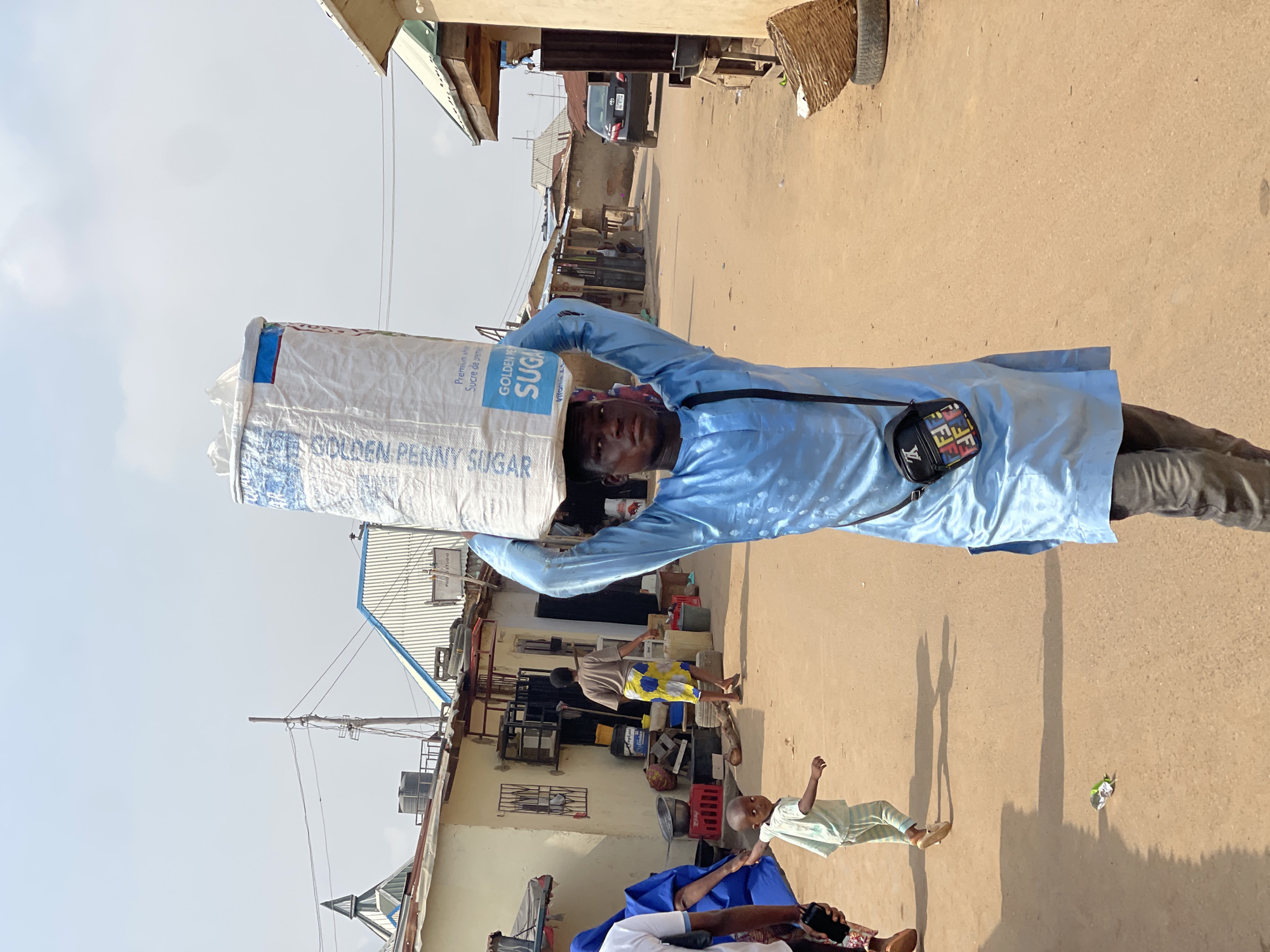The image depicts a daytime street scene in Kuchingoro, Nigeria. A man, wearing a light blue long-sleeved garment and dark trousers, is prominently featured, carrying a large white sack on his head. The sack is labeled "GOLDEN PENNY SUGAR" and also shows text like "Premium wh" and "Sucre de premie". A black shoulder bag with colorful "FIF" lettering is slung across his chest.
The man stands on an unpaved, dusty road, which recedes into the background. Buildings with corrugated metal roofs line both sides of the street, suggesting a residential or commercial area. In the background, other individuals are visible, including another adult standing near a building and a small child in striped clothing standing on the ground. A vehicle is partially visible further down the street. Long shadows indicate bright sunlight, likely late morning or afternoon. The scene conveys an everyday activity, possibly a vendor or someone transporting goods.