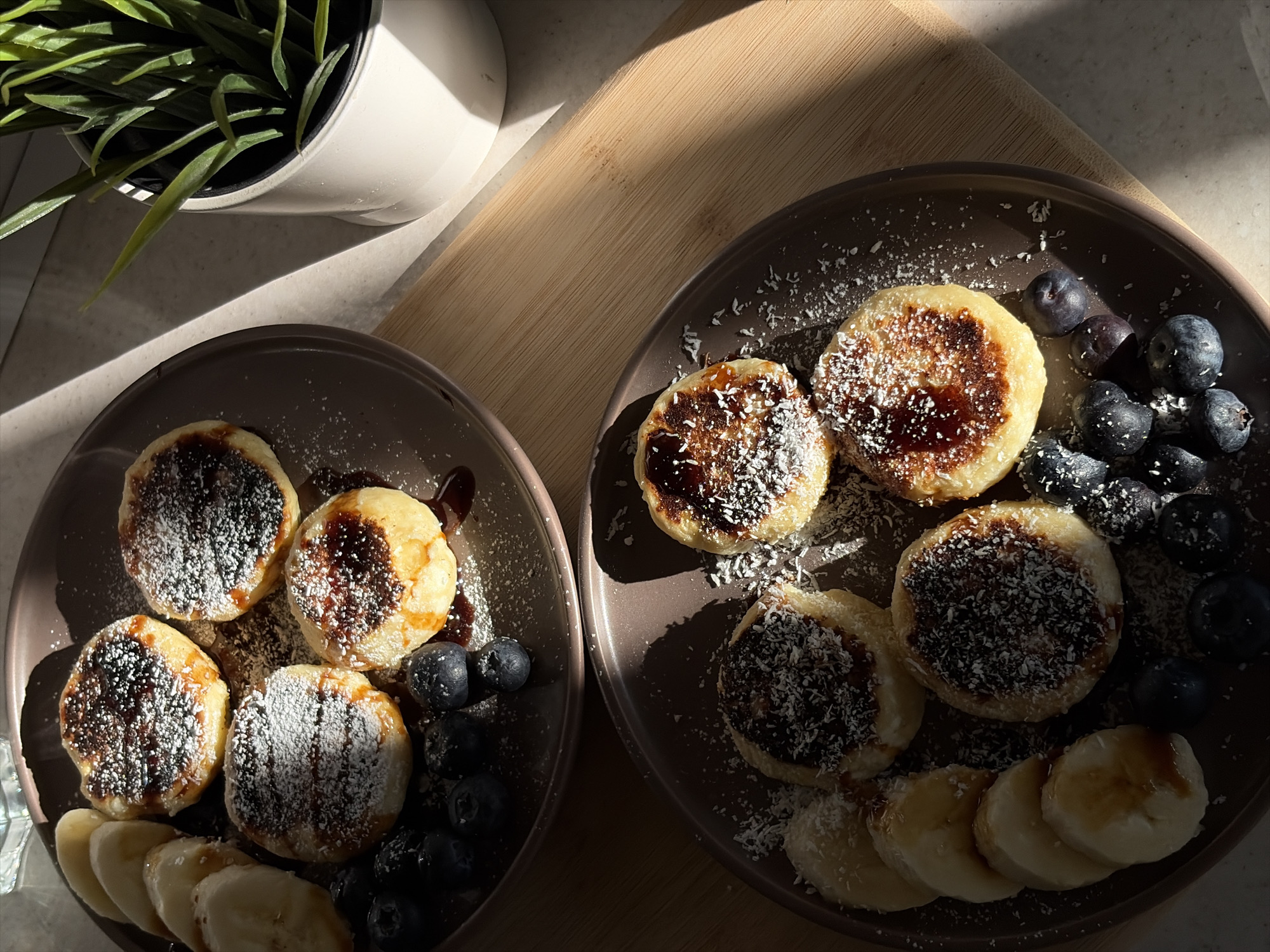Two brown plates of food are situated on a wooden surface, likely a countertop or table. The plate on the left contains four small, round pancakes, drizzled with a dark syrup and dusted with a white powder, possibly powdered sugar or coconut. Sliced bananas and blueberries are arranged around the pancakes. The plate on the right mirrors this setup, containing four similar pancakes with syrup and white powder, along with blueberries and banana slices. To the left, a white pot holding a small green plant adds a natural element to the scene. The scene is illuminated by natural sunlight, creating contrasting areas of light and shadow. The location is Chaikovsky, Russia.
