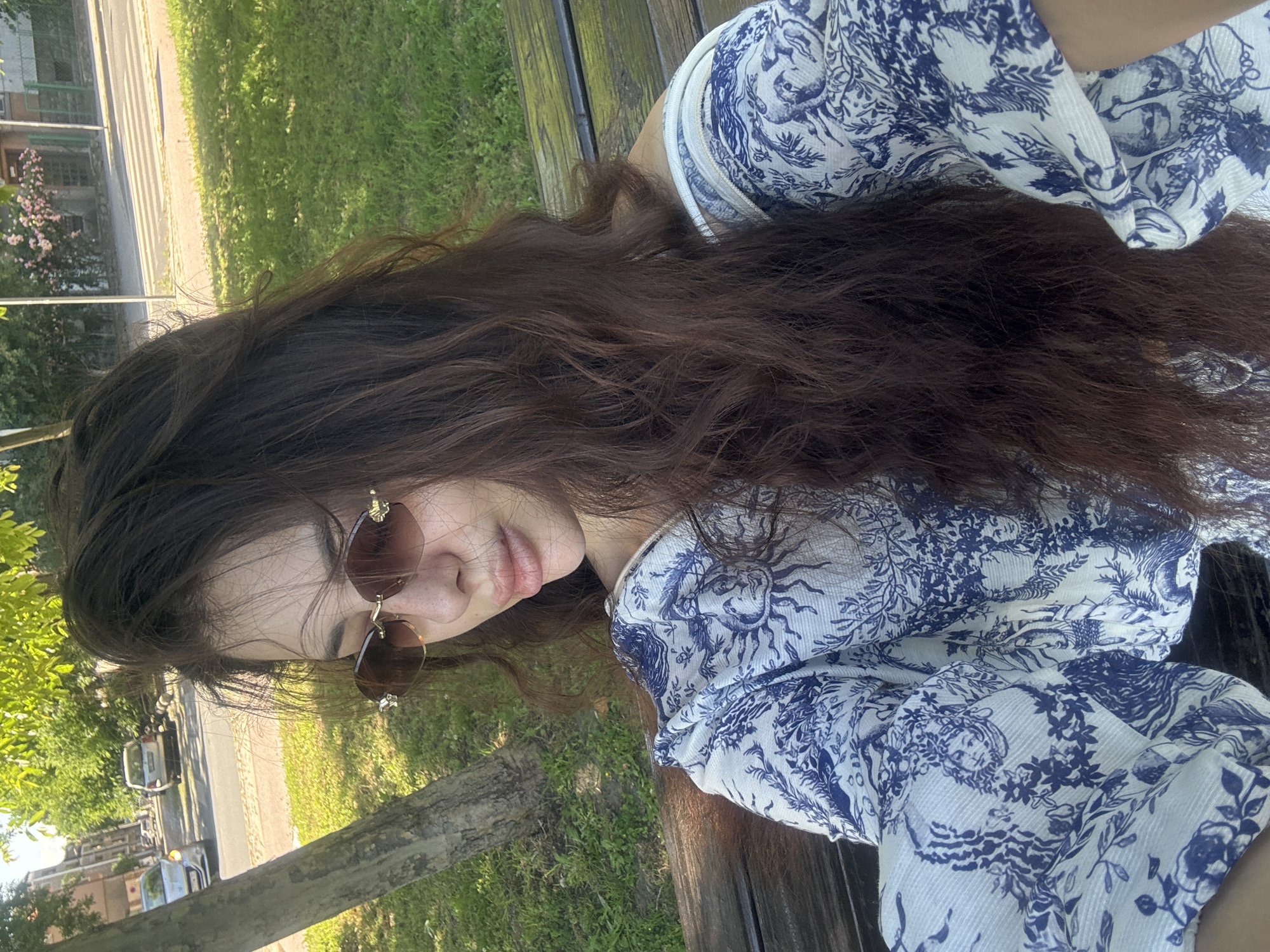 A young woman is sitting outside in Plovdiv, Bulgaria. She has long brown hair and is wearing sunglasses and a blue and white patterned shirt. She is sitting on a wooden surface, possibly a bench. In the background, there is green grass, trees, and a street with parked cars. A building is visible in the distance. The image appears to be taken during the daytime.