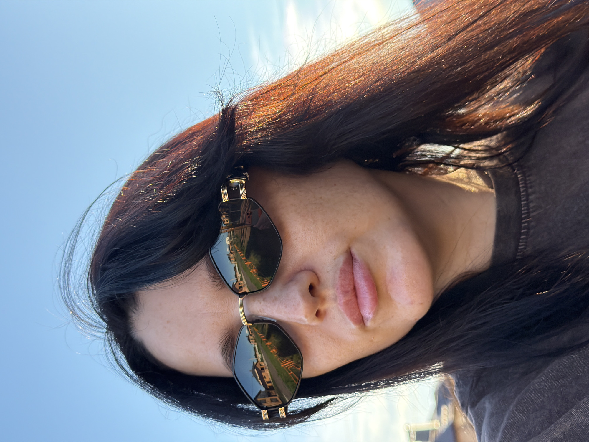 A close-up, outdoor, eye-level, medium shot captures a woman with dark hair and fair skin wearing sunglasses. The woman's face is turned slightly to the left, with her lips slightly parted. Her hair is dark and falls around her face and shoulders, with strands catching the sunlight. The sunglasses have a diamond-shaped lens with a black frame and gold accents. The reflection in the sunglasses shows a street lined with houses and an orange fence, suggesting a suburban or rural setting. The background consists of a clear blue sky. The image appears to be taken in the city of Shmidtovo, Russia, likely during daylight hours given the sunlight.