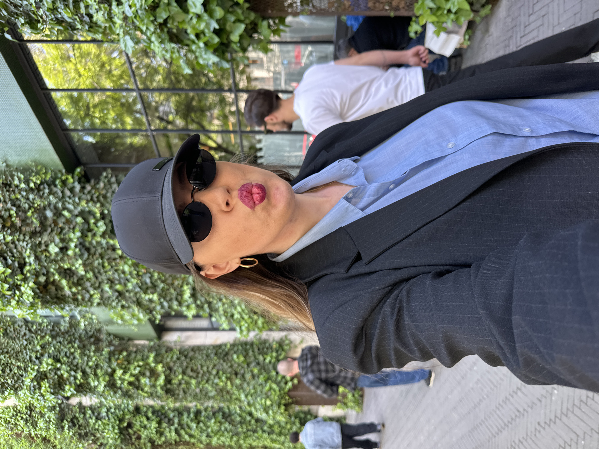 A woman wearing sunglasses and a baseball cap poses for a selfie. She has applied dark lipstick and is pouting her lips. She is wearing a light blue button-down shirt and a dark pinstripe blazer. She is standing in front of a building with a wall covered in ivy. Other people can be seen walking on the sidewalk behind her. Based on the ivy-covered wall and architectural style, the location is likely in Tbilisi, Georgia.