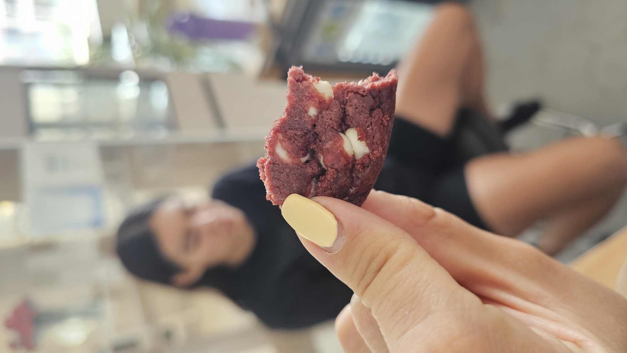 A close-up, shallow depth-of-field image features a hand holding a bitten red velvet cookie with white chocolate chips. The cookie, dominant in the foreground, is a vibrant crimson, displaying its crumbly texture and generous inclusion of irregularly shaped white chocolate pieces. A portion of the cookie has been bitten off, revealing its interior. The hand holding the cookie is lightly tanned, and the fingernail of the pointer finger is painted a solid, pale yellow.
The background is heavily blurred, creating a bokeh effect that draws focus to the cookie. In the middle ground, a person is visible lying down in a relaxed posture, with their legs slightly elevated. They are wearing dark clothing, appearing to be a dark top and shorts. The distant background consists of indistinct, out-of-focus shapes and light sources, suggesting an indoor setting with shelves or furniture. The lighting appears soft and natural, indicating daytime. No visible text can be deciphered in the blurred background. The scene suggests a casual, leisurely moment, possibly taking place in Puebla, Mexico.